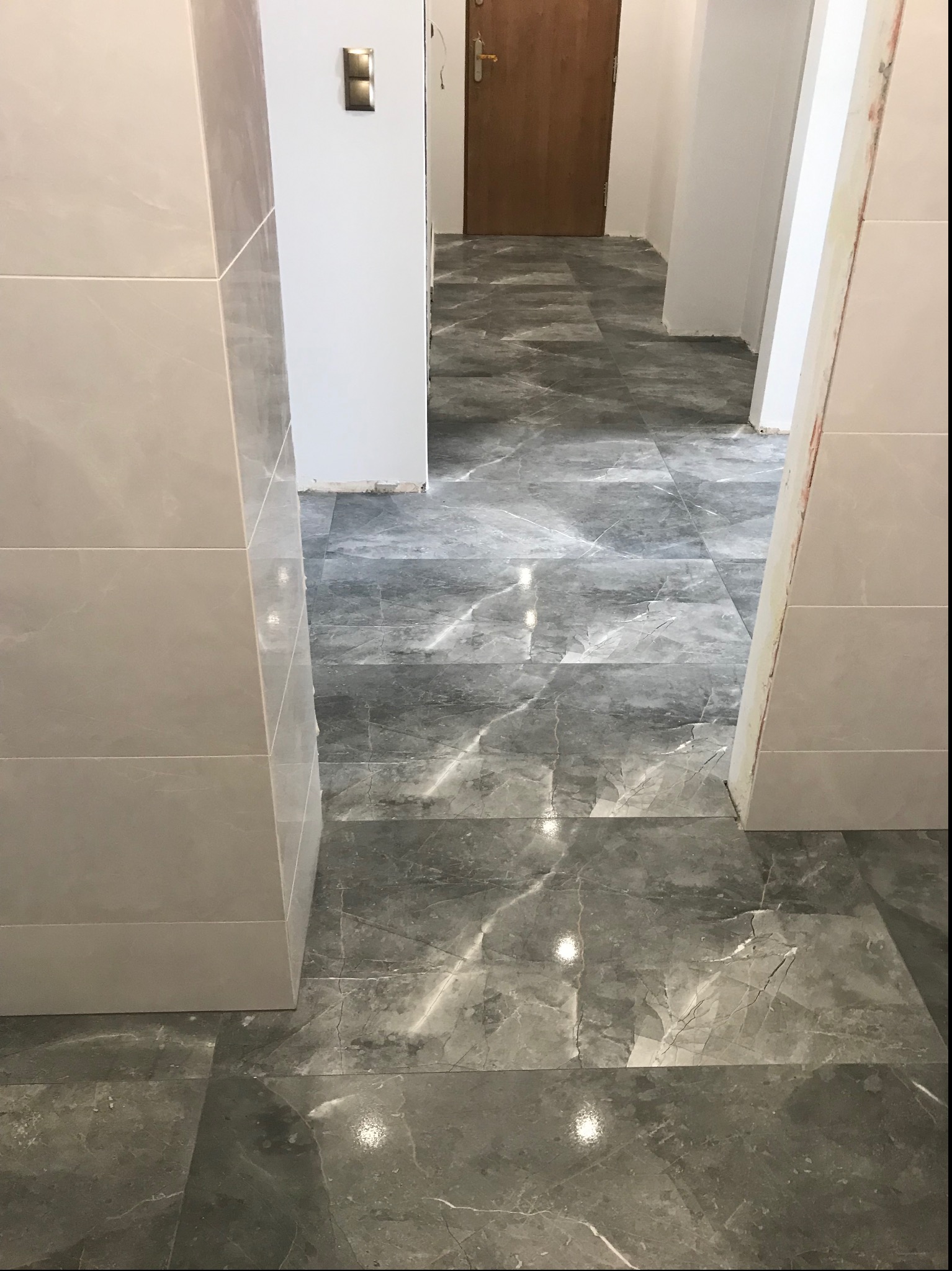 Ułożona szara marmurkowa podłoga z płytek w korytarzu, widoczne jasne płytki na ścianie i fragment drewnianych drzwi w tle.