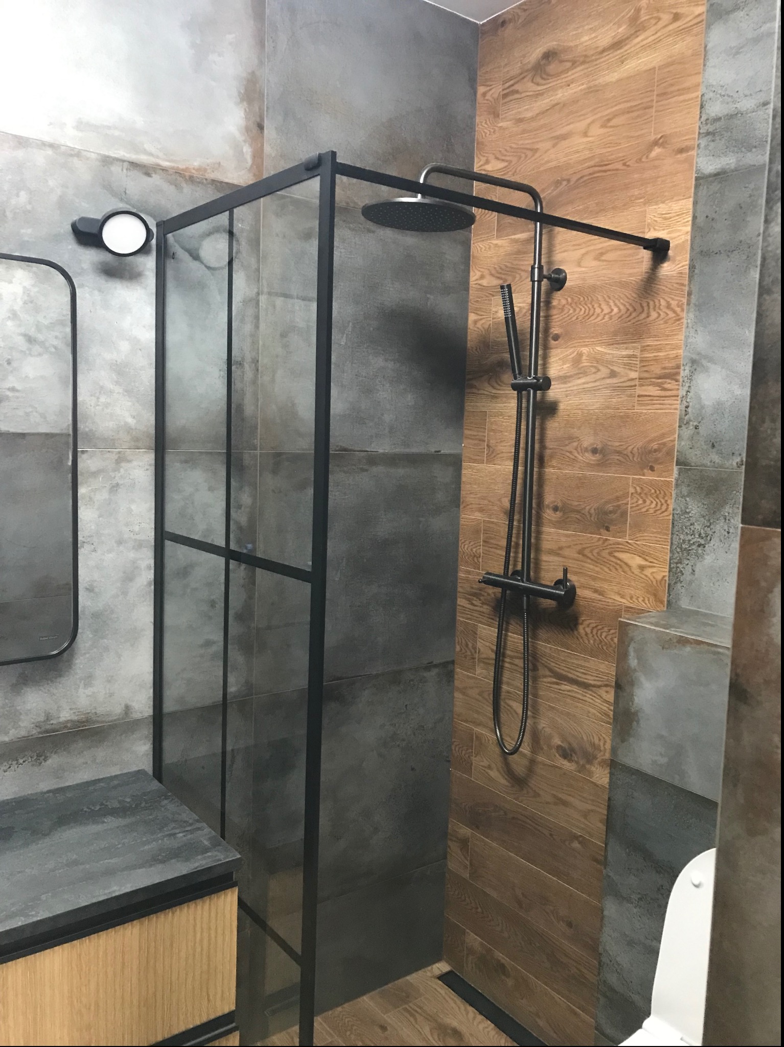 Nowoczesna kabina prysznicowa z czarną ramą i deszczownicą na tle płytek imitujących drewno i szary beton, oświetlona punktowym światłem.