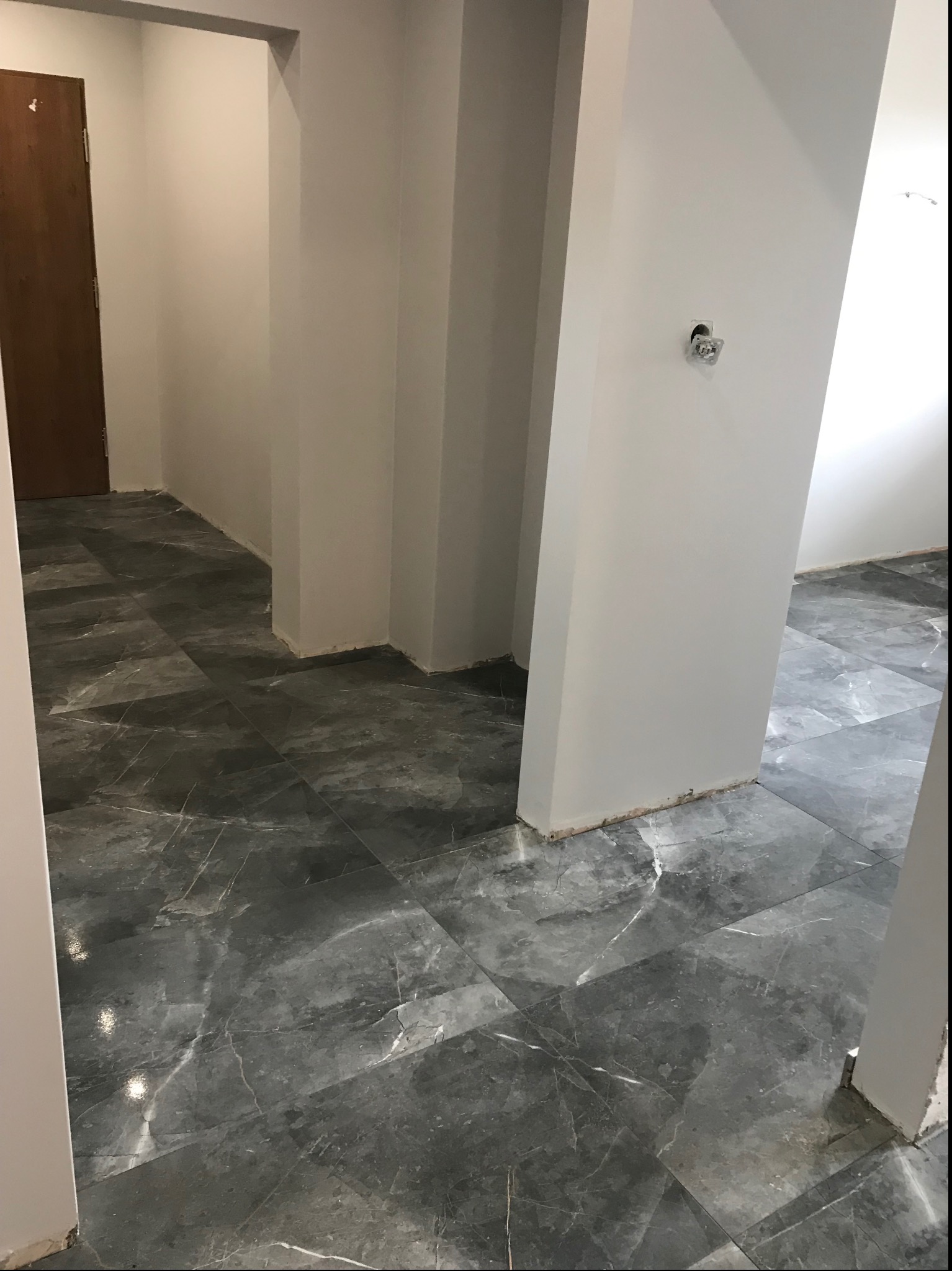Korytarz w trakcie remontu, z położoną ciemnoszarą marmurkową podłogą z płytek, świeżo pomalowanymi na biało ścianami i widoczną puszką elektryczną bez osprzętu.