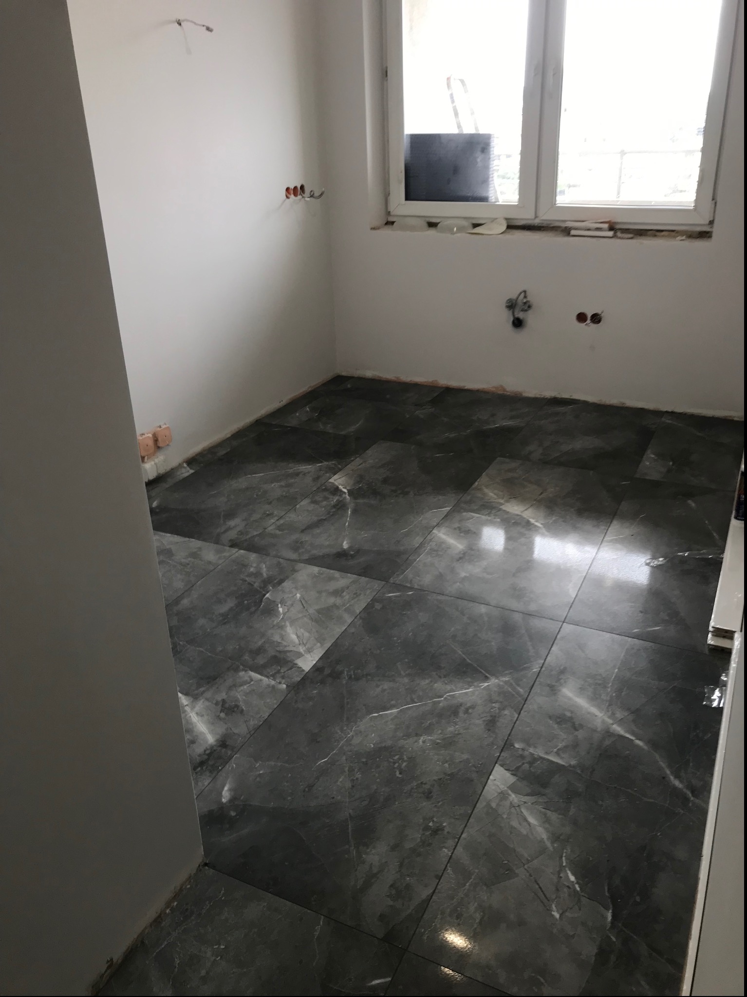 Ułożone, ciemnoszare płytki imitujące marmur na podłodze w łazience, z widocznymi puszkami elektrycznymi w ścianie i oknem w tle.