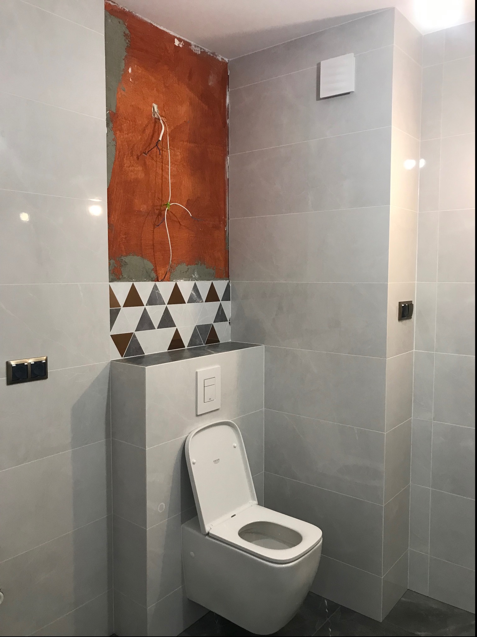 Świeżo wykafelkowana łazienka z częściowo przygotowaną ścianą do dalszych prac wykończeniowych, widoczne kable elektryczne i dekoracyjna mozaika z trójkątów.
