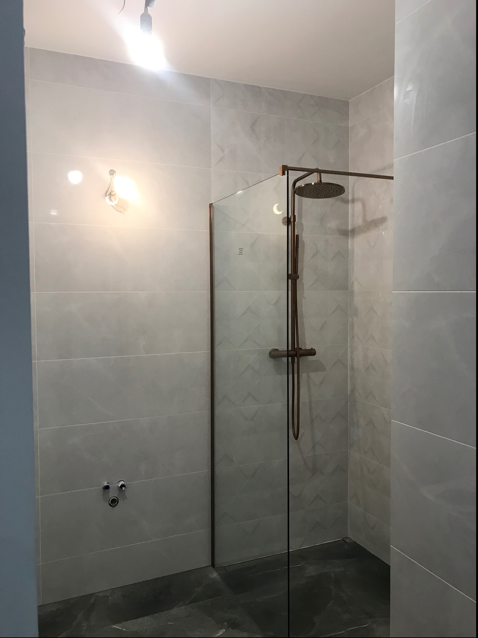 Nowoczesna kabina prysznicowa typu walk-in z miedzianą armaturą na tle jasnoszarych płytek w geometryczny wzór, widoczne przygotowane przyłącza wodne.