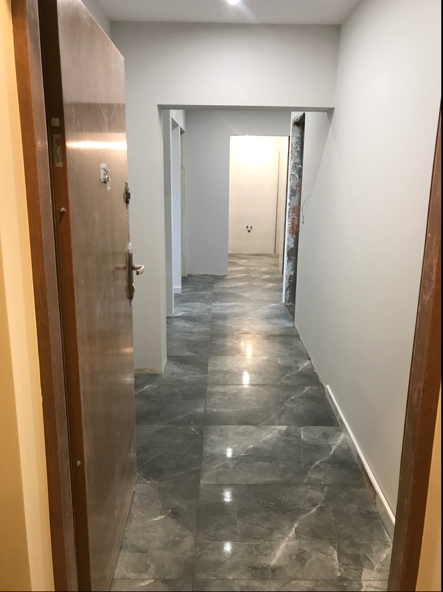 Korytarz po remoncie z szarymi płytkami imitującymi marmur na podłodze, świeżo pomalowane ściany na szaro, widok przez otwarte drzwi wejściowe na łazienkę w głębi.