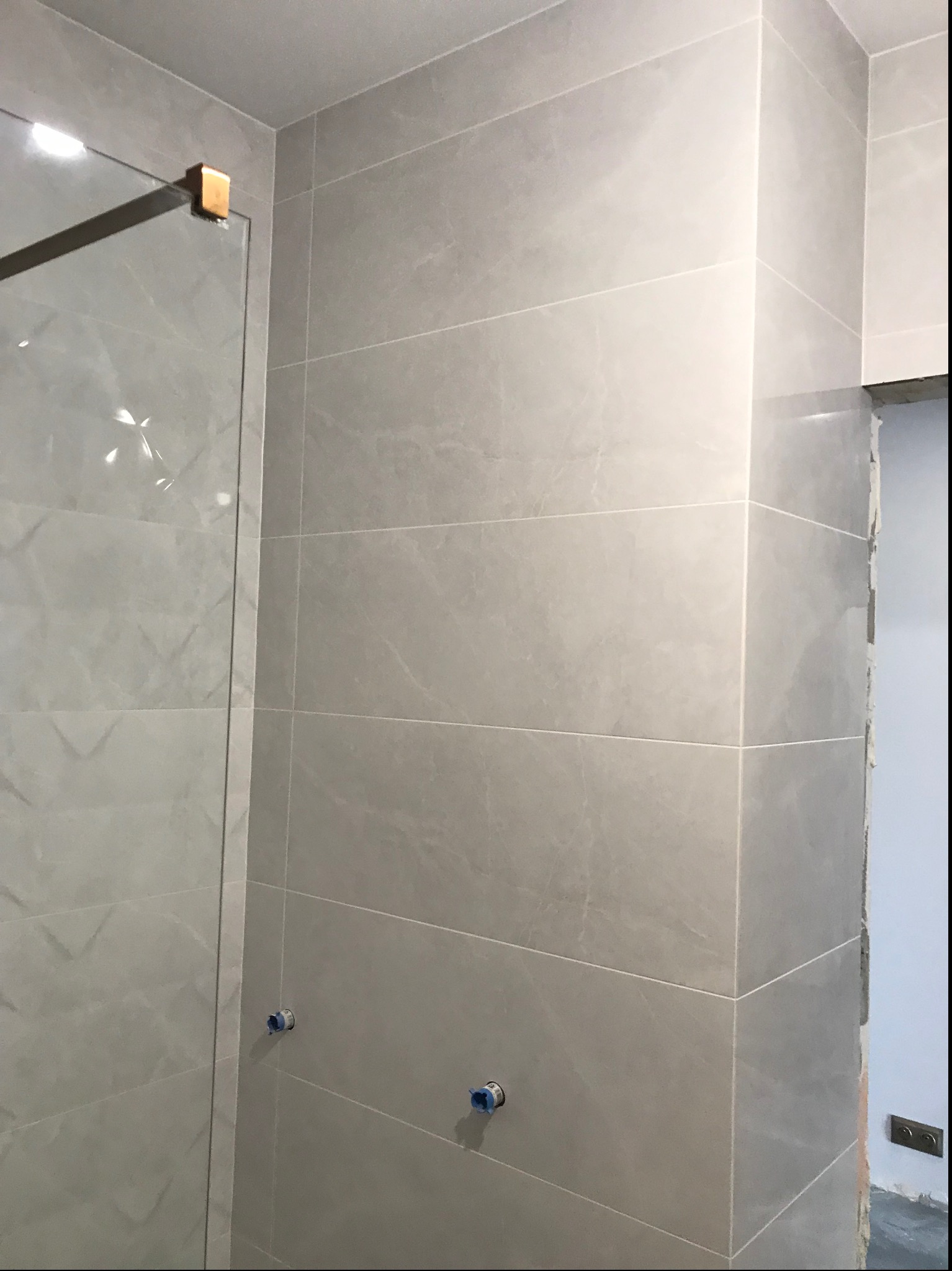 Świeżo wykafelkowana ściana łazienki w odcieniach szarości z widocznymi przyłączami wodnymi i fragmentem kabiny prysznicowej z elementami w kolorze mosiądzu.