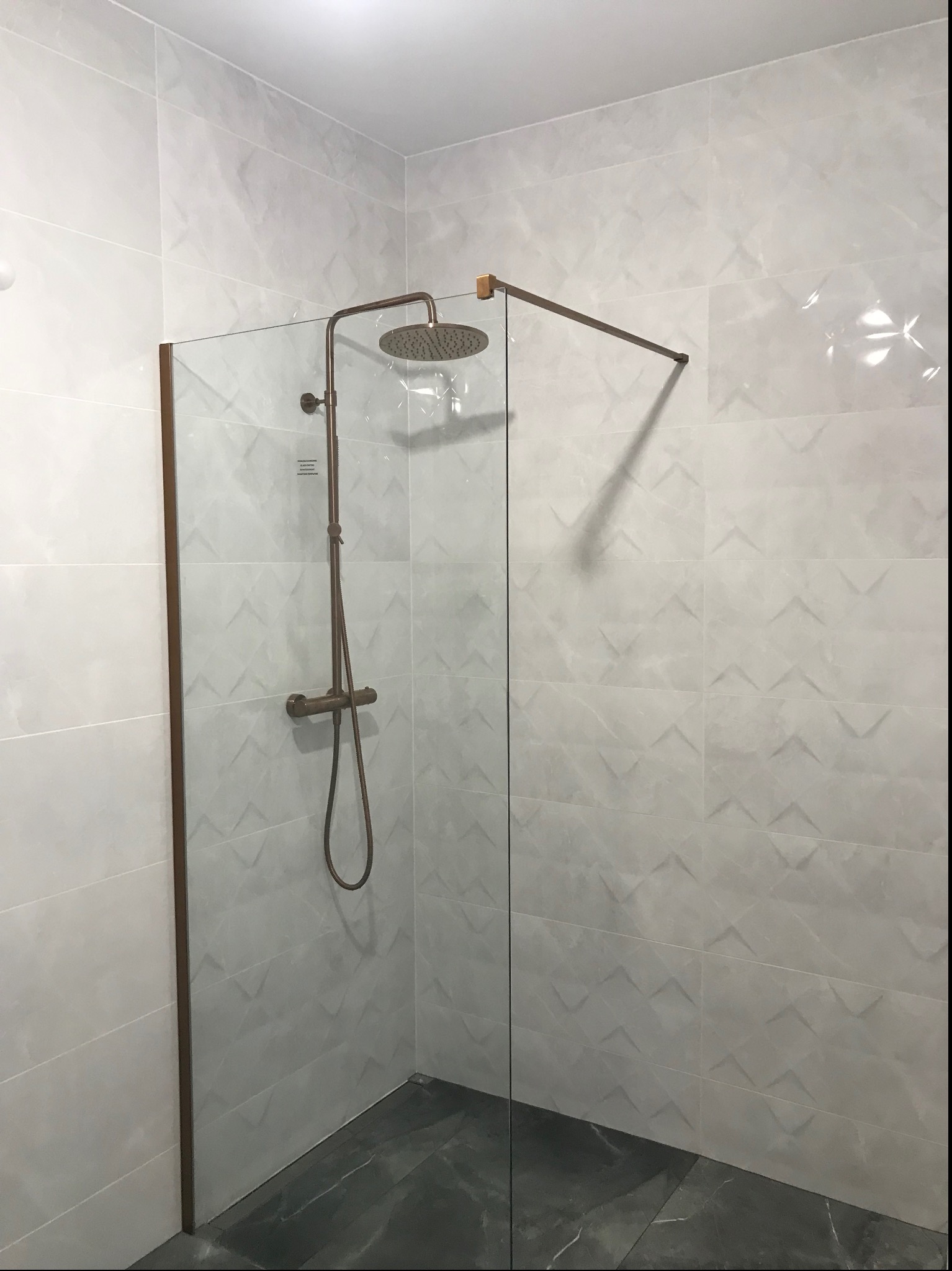 Wykończona kabina prysznicowa typu walk-in z miedzianą armaturą i płytkami w odcieniach szarości z subtelnym wzorem geometrycznym na ścianach, oraz ciemną marmurową posadzką.