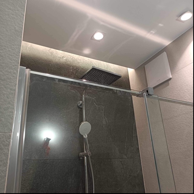 Wykończona kabina prysznicowa z deszczownicą i słuchawką, szare płytki imitujące kamień, oświetlenie punktowe i wentylator sufitowy w nowoczesnej łazience.