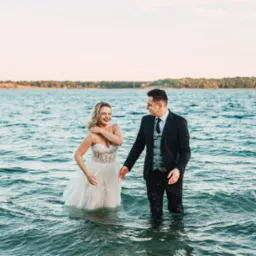 Uśmiechnięta panna młoda w koronkowej sukni i pan młody w garniturze stoją w wodzie jeziora podczas sesji ślubnej na tle zalesionego brzegu o zachodzie słońca.