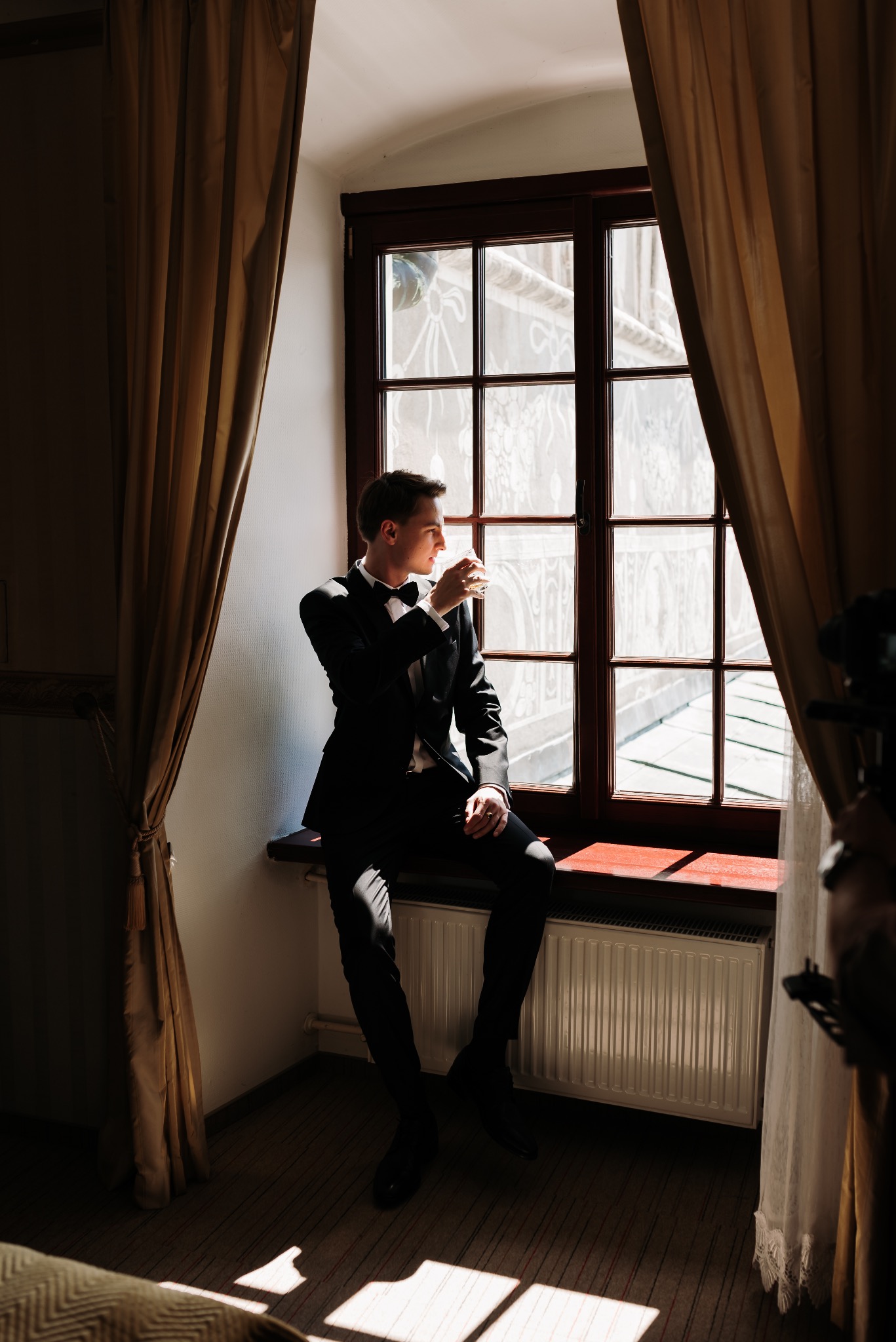 Elegancki mężczyzna w czarnym smokingu z muszką, siedzący na parapecie okna z szprosami, popijający drink i spoglądający na zewnątrz, oświetlony ciepłym światłem słonecznym wpadającym przez okno...