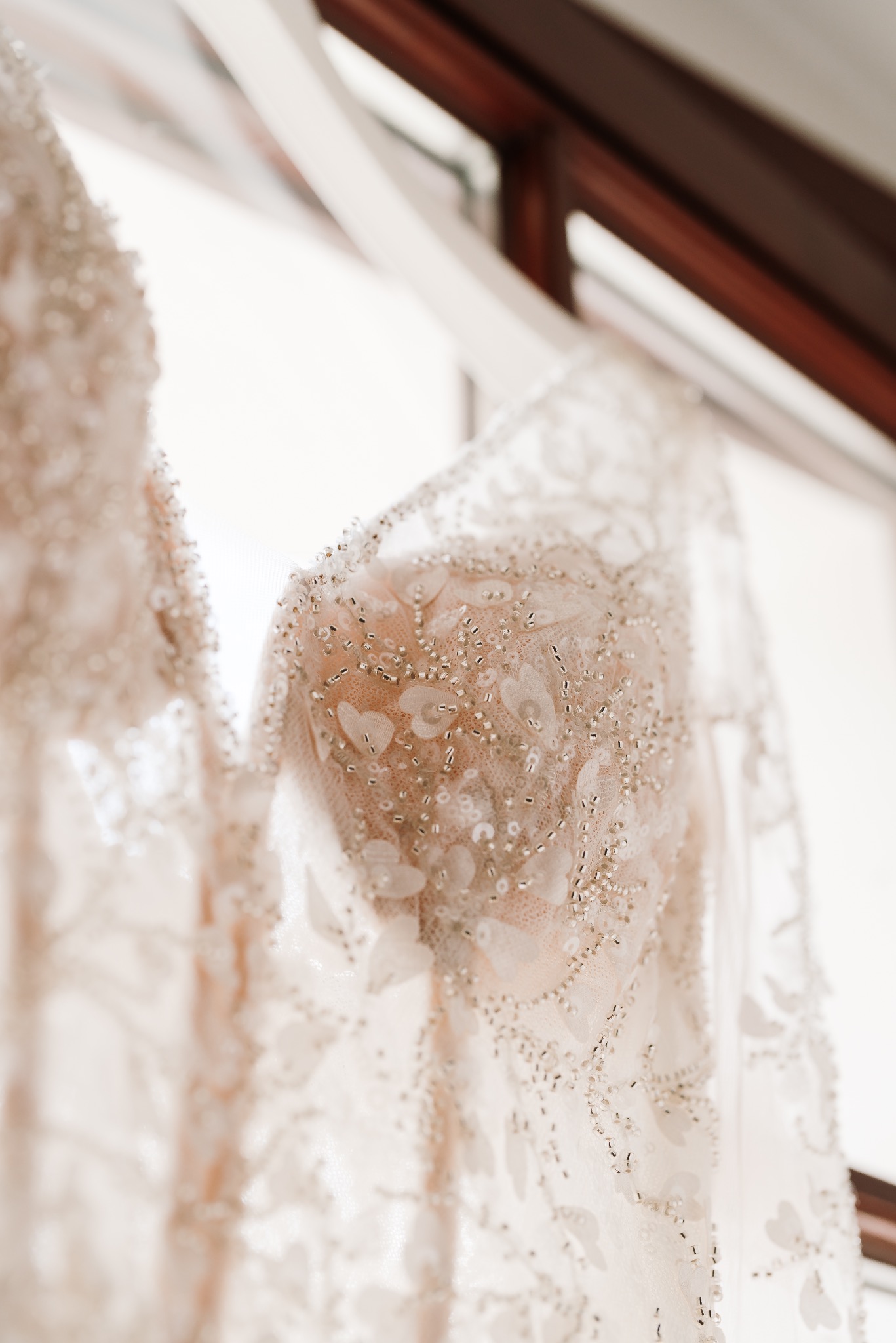 Delikatne zbliżenie na górną część sukni ślubnej z koronkowymi aplikacjami i drobnymi koralikami, wiszącej przy oknie.