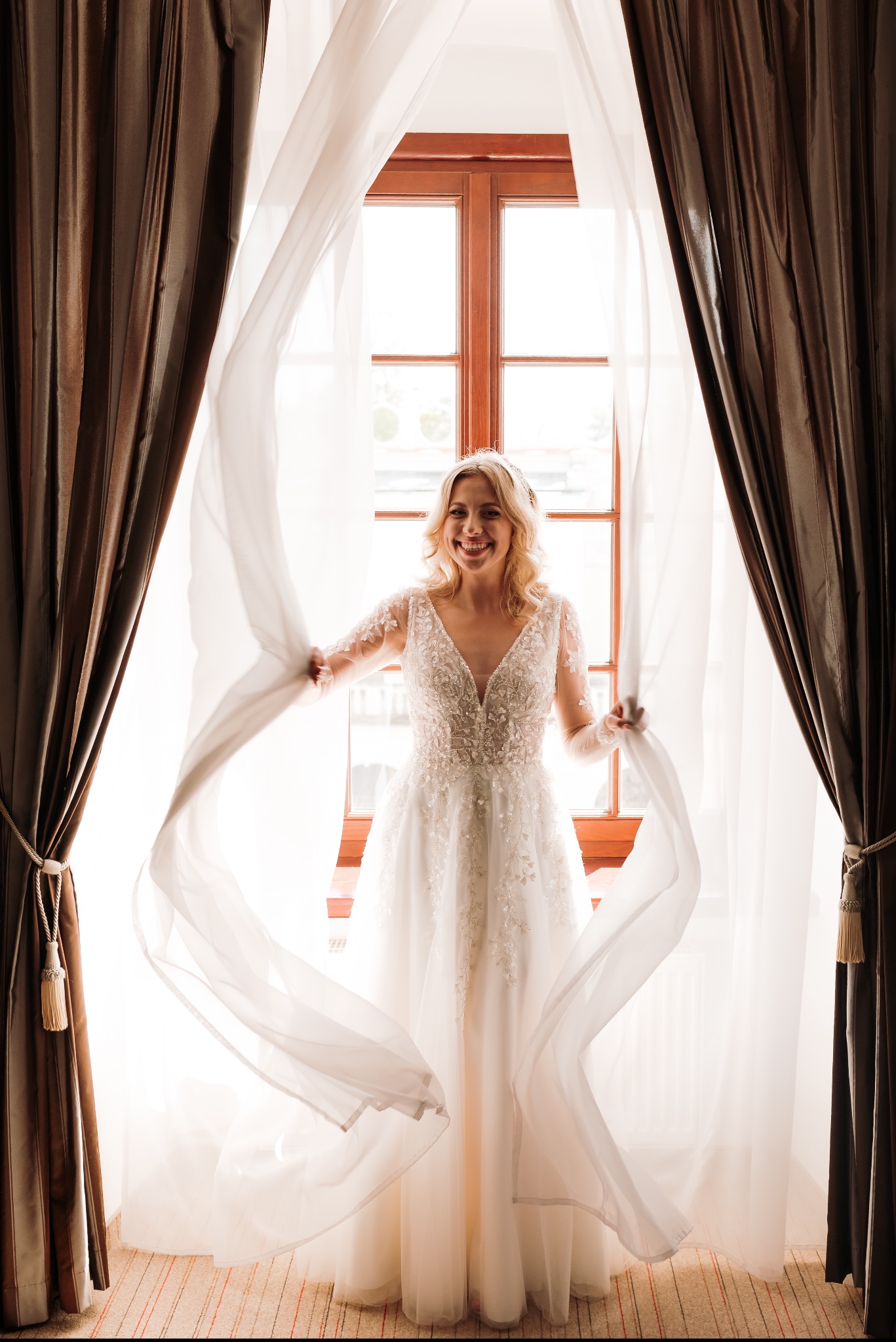 Uśmiechnięta panna młoda w koronkowej sukni, stojąca przy oknie z powiewającymi, półprzezroczystymi zasłonami