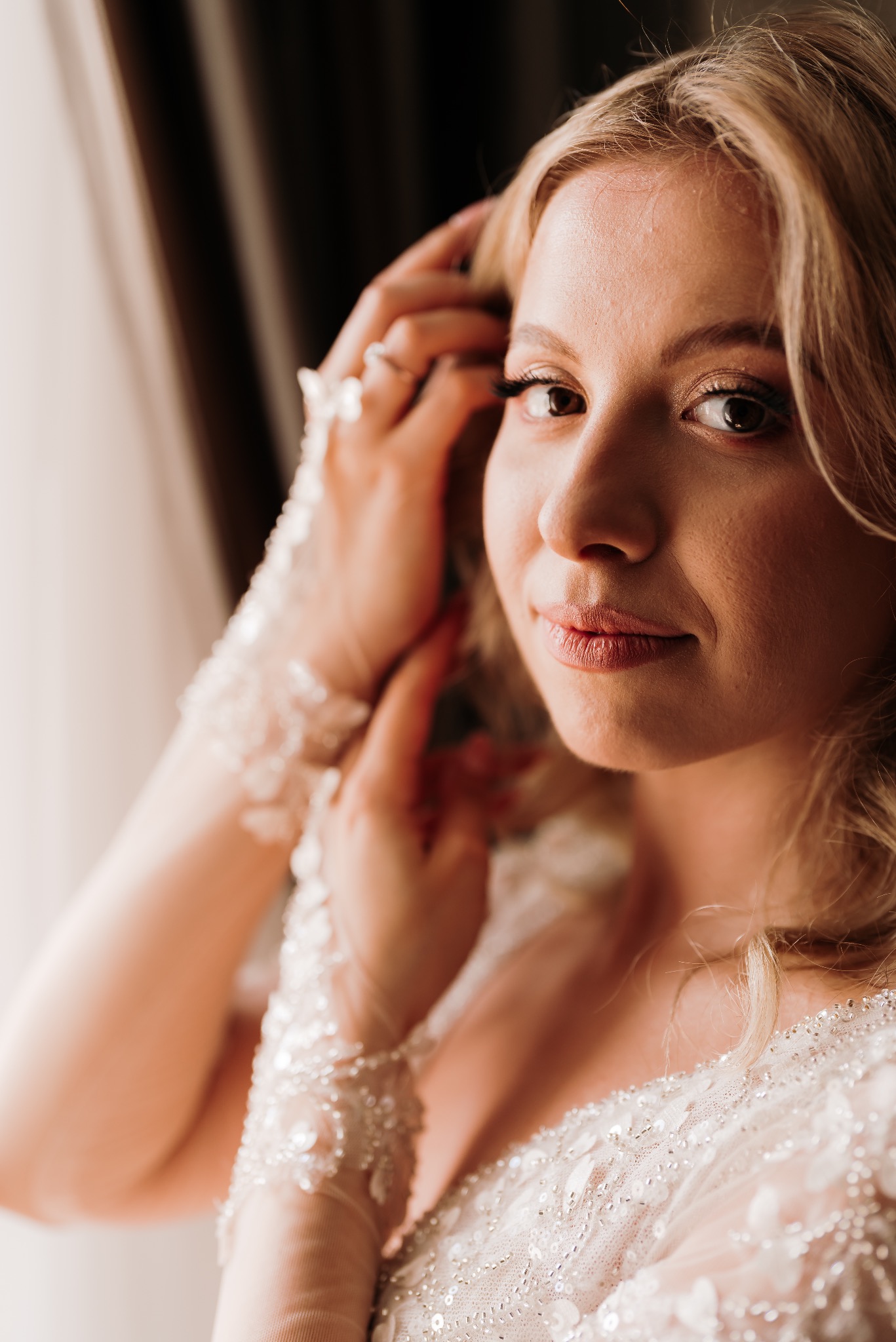 Panna młoda w sukni ślubnej z koronkowymi rękawami, poprawia włosy przed ceremonią, delikatny makijaż i pierścionek zaręczynowy.