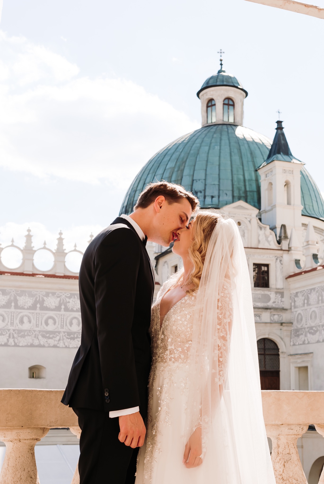 Pocałunek nowożeńców na tle barokowej architektury z kopułą, panna młoda w sukni z kryształkami i długim welonem, pan młody w czarnym smokingu.