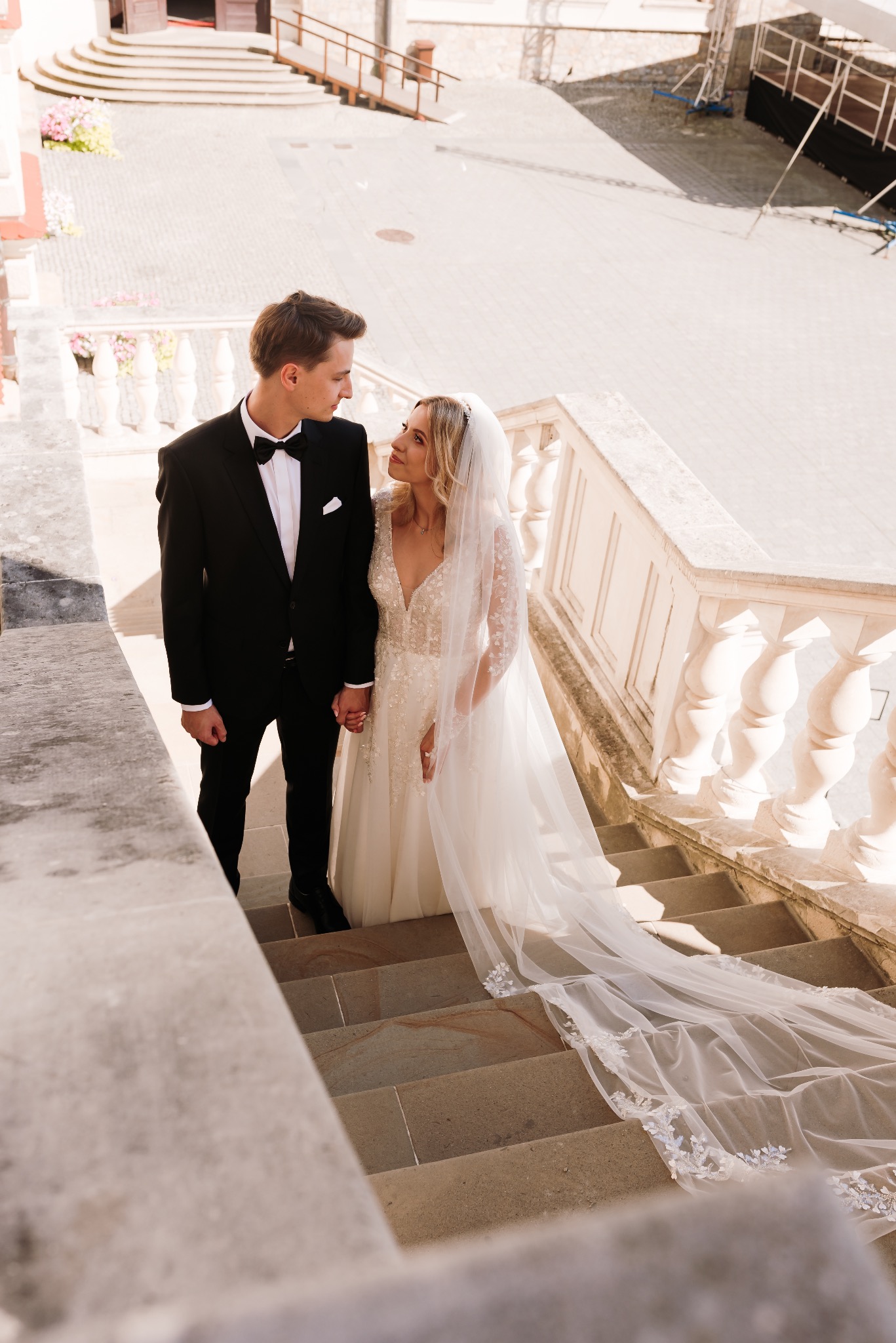 Para młoda trzymająca się za ręce na kamiennych schodach z balustradą, panna młoda w sukni z długim, koronkowym welonem.