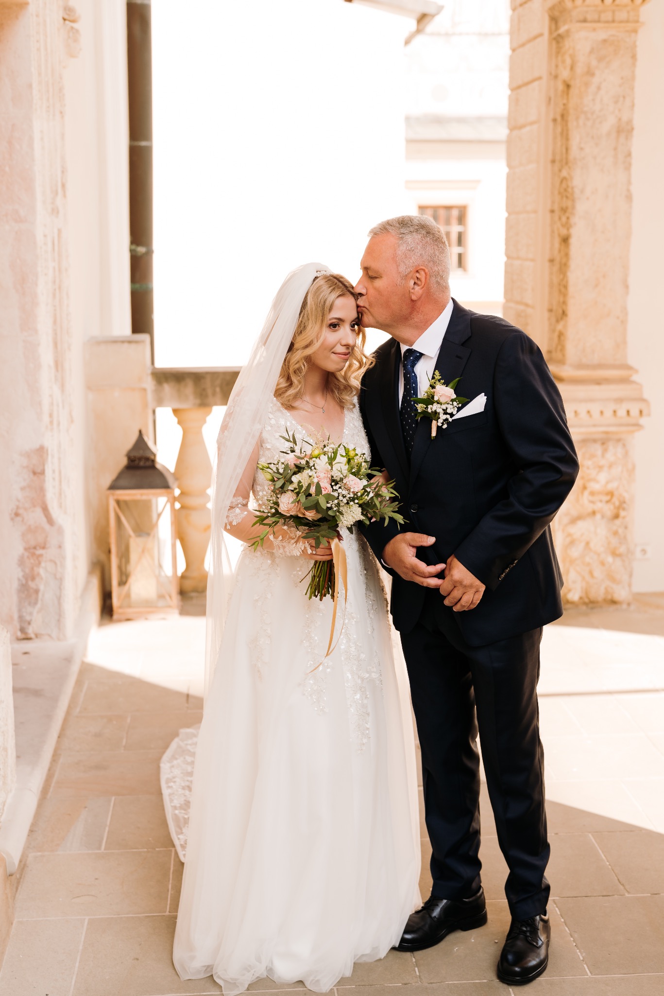 Panna młoda w białej sukni z bukietem kwiatów, całowana w czoło przez mężczyznę w garniturze na tle kolumn i kamiennej posadzki.