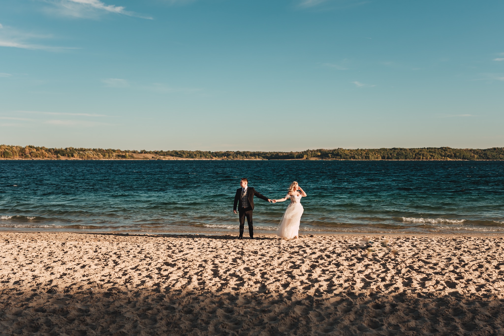 Para młoda trzymająca się za ręce na piaszczystej plaży nad jeziorem w słoneczny dzień. W tle widoczny zalesiony brzeg i błękitne niebo z lekkimi obłokami.