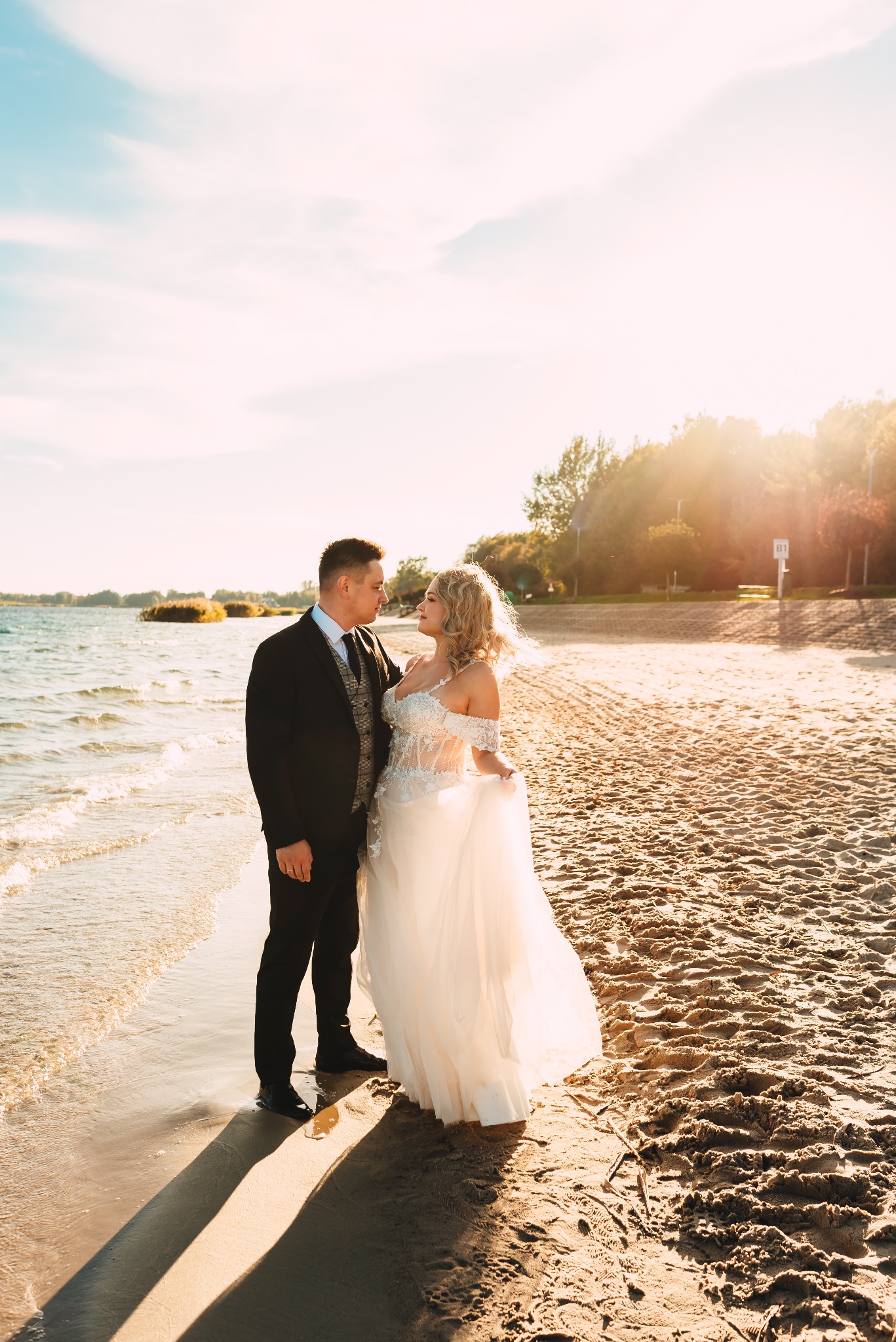 Para młoda w eleganckich strojach stoi na piaszczystej plaży, spoglądając na siebie w promieniach zachodzącego słońca, z widokiem na jezioro i drzewa w tle.