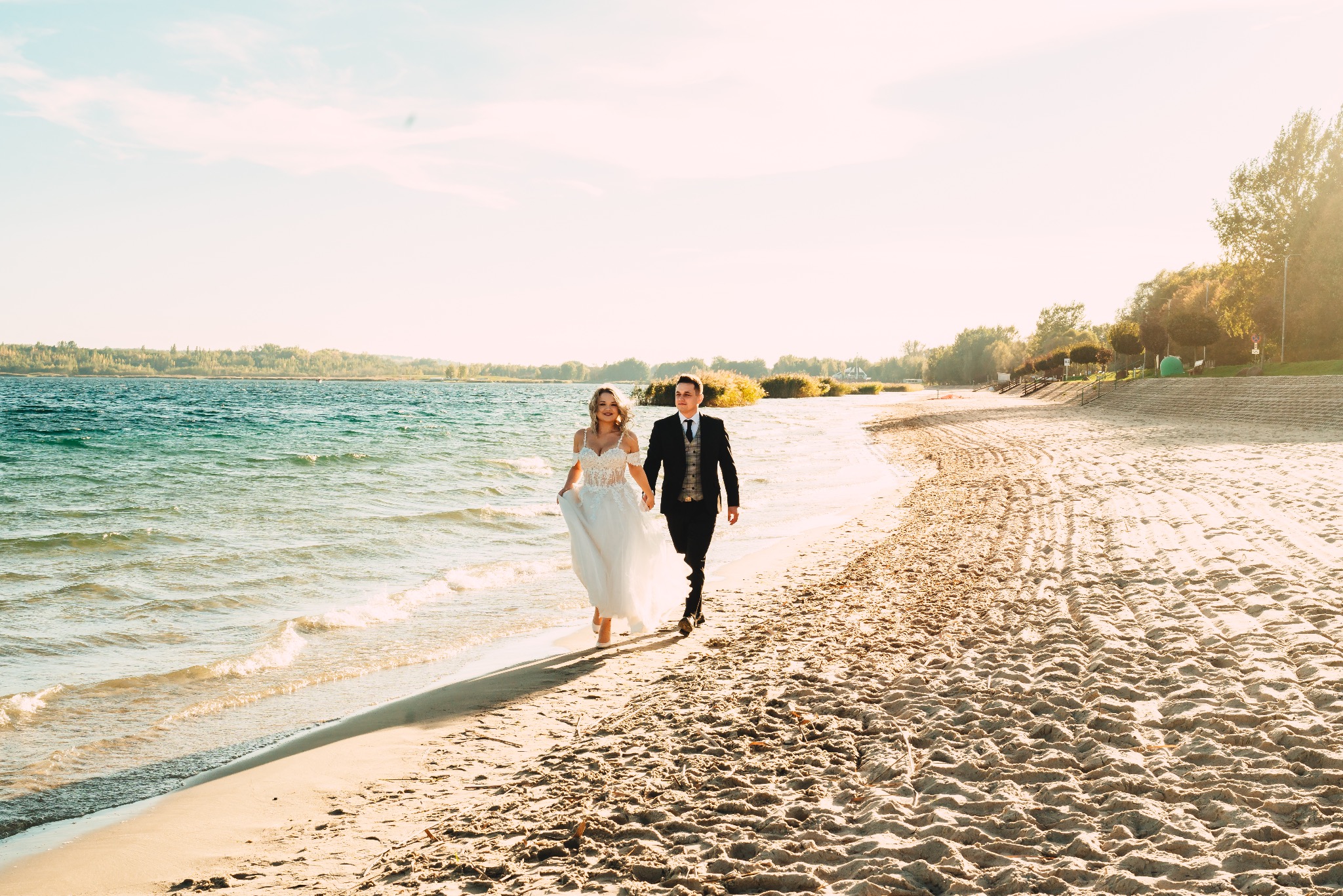 Para młoda spaceruje brzegiem morza w promieniach zachodzącego słońca, panna młoda w koronkowej sukni, pan młody w eleganckim garniturze.