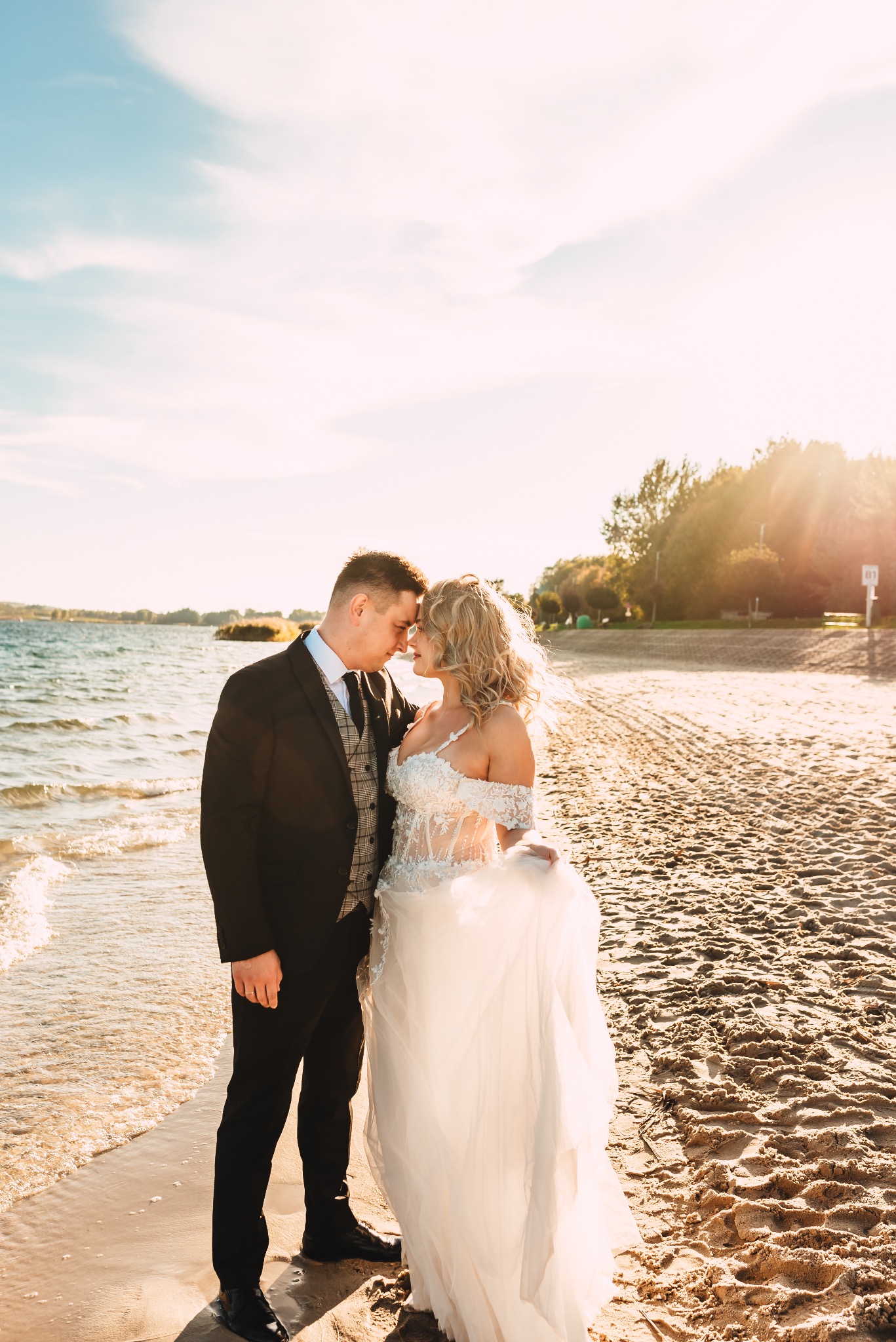 Para młoda, stojąca na piaszczystej plaży nad brzegiem jeziora, w promieniach zachodzącego słońca, panna młoda w koronkowej sukni z odkrytymi ramionami i blond lokami, pan młody w czarnym...