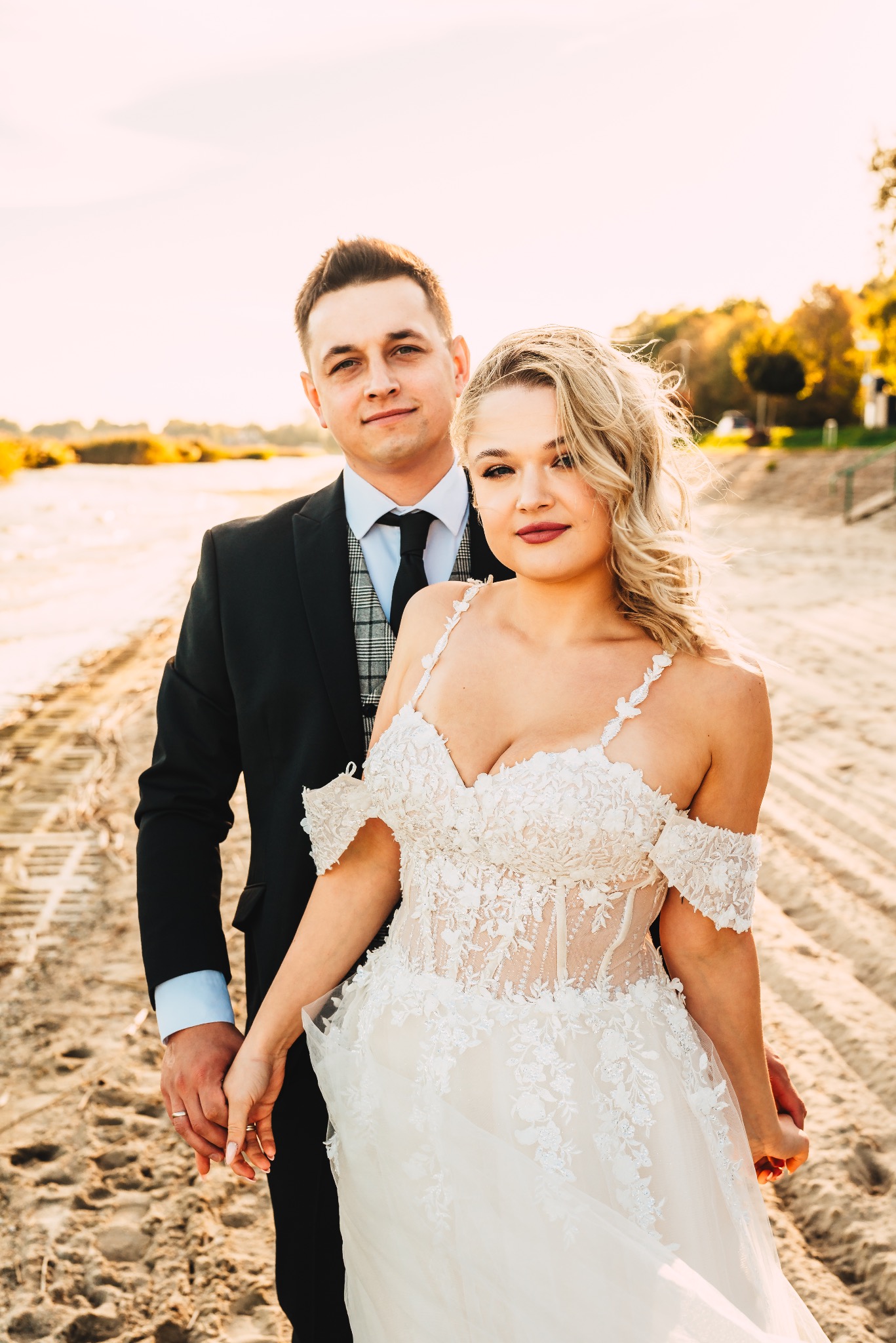 Para młoda trzymająca się za ręce na plaży w sukni ślubnej z koronkowymi aplikacjami i garniturze z kraciastą kamizelką, w tle drzewa i piasek z wyraźnymi śladami opon.