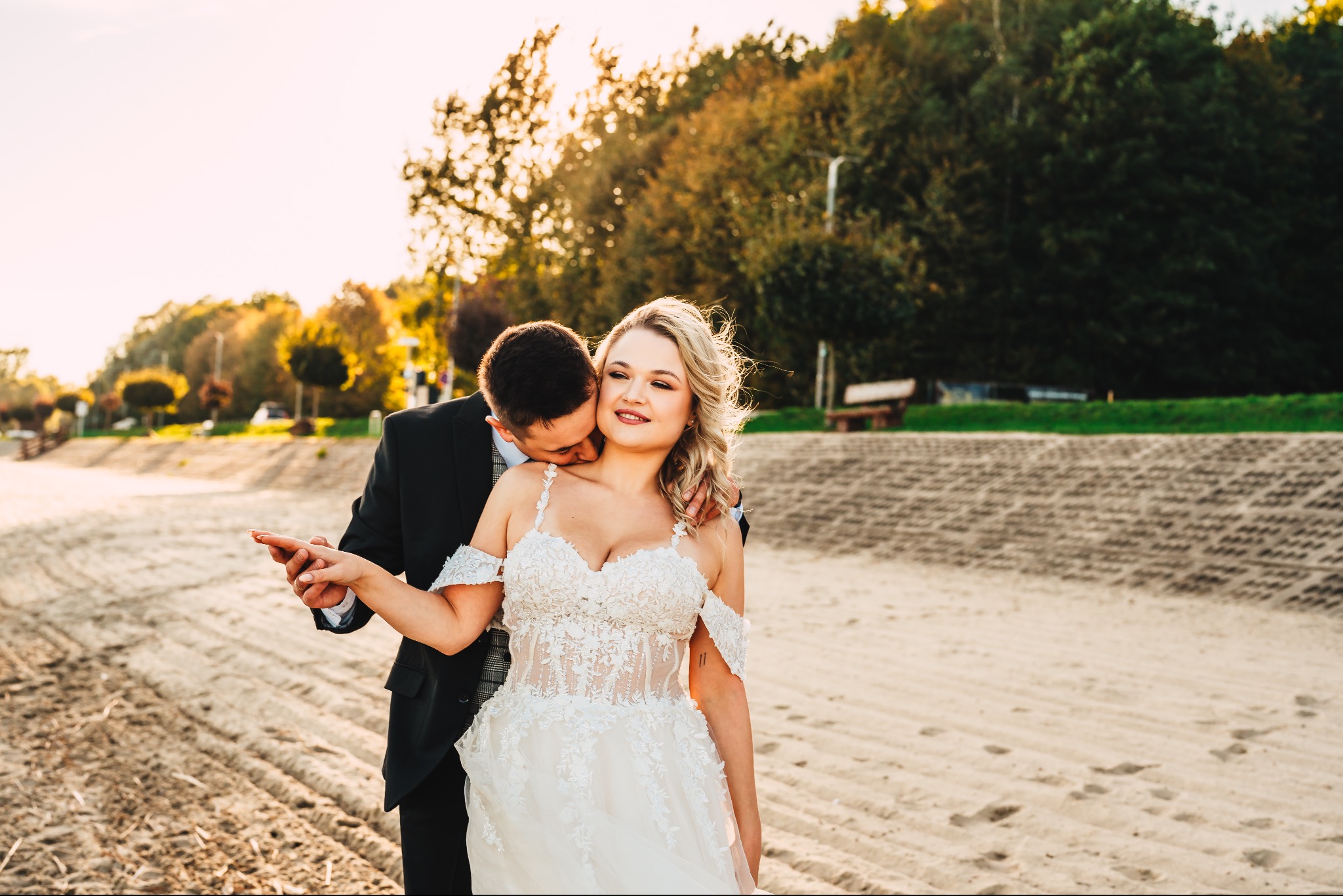 Para młoda na plaży w jesiennym słońcu: Panna młoda w koronkowej sukni, pan młody w czarnym garniturze całuje ją w szyję, trzymając ją za rękę.
