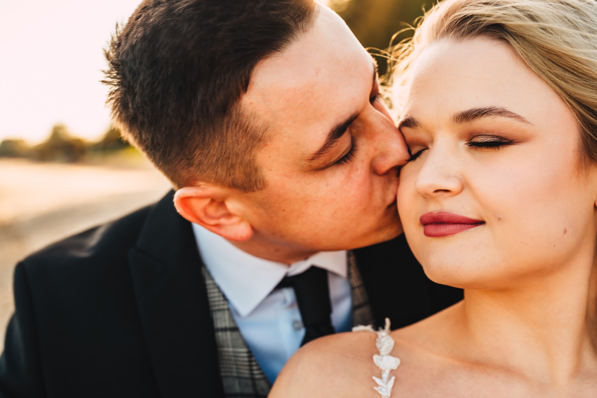 Pan młody w czarnym garniturze całuje pannę młodą w powiekę, panna młoda ma zamknięte oczy, widoczny delikatny makijaż i biała sukienka z kwiatowymi aplikacjami.