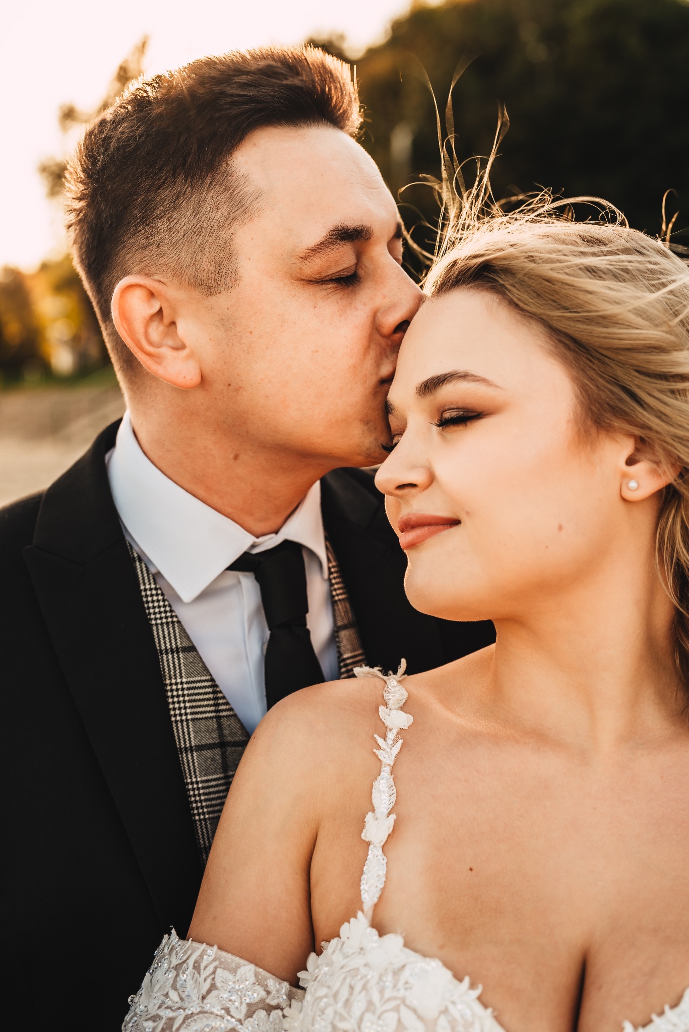 Panna młoda w sukni ślubnej z dekorowanymi ramiączkami, z zamkniętymi oczami, całowana w czoło przez pana młodego w czarnym garniturze z wzorzystą kamizelką. Delikatne promienie słońca muskają...