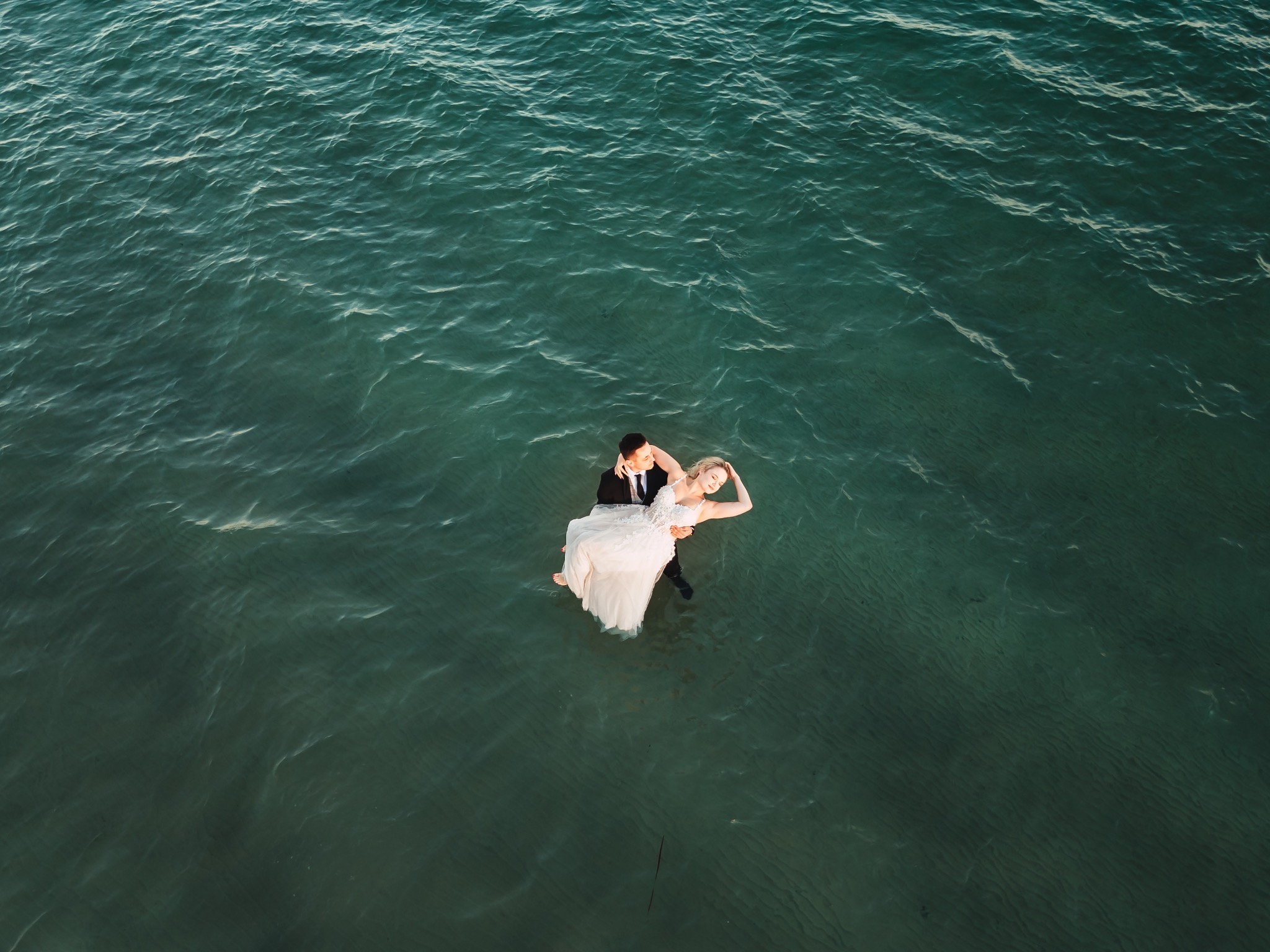 Mężczyzna w smokingu trzyma pannę młodą w białej sukni na tle turkusowej wody, widok z góry.