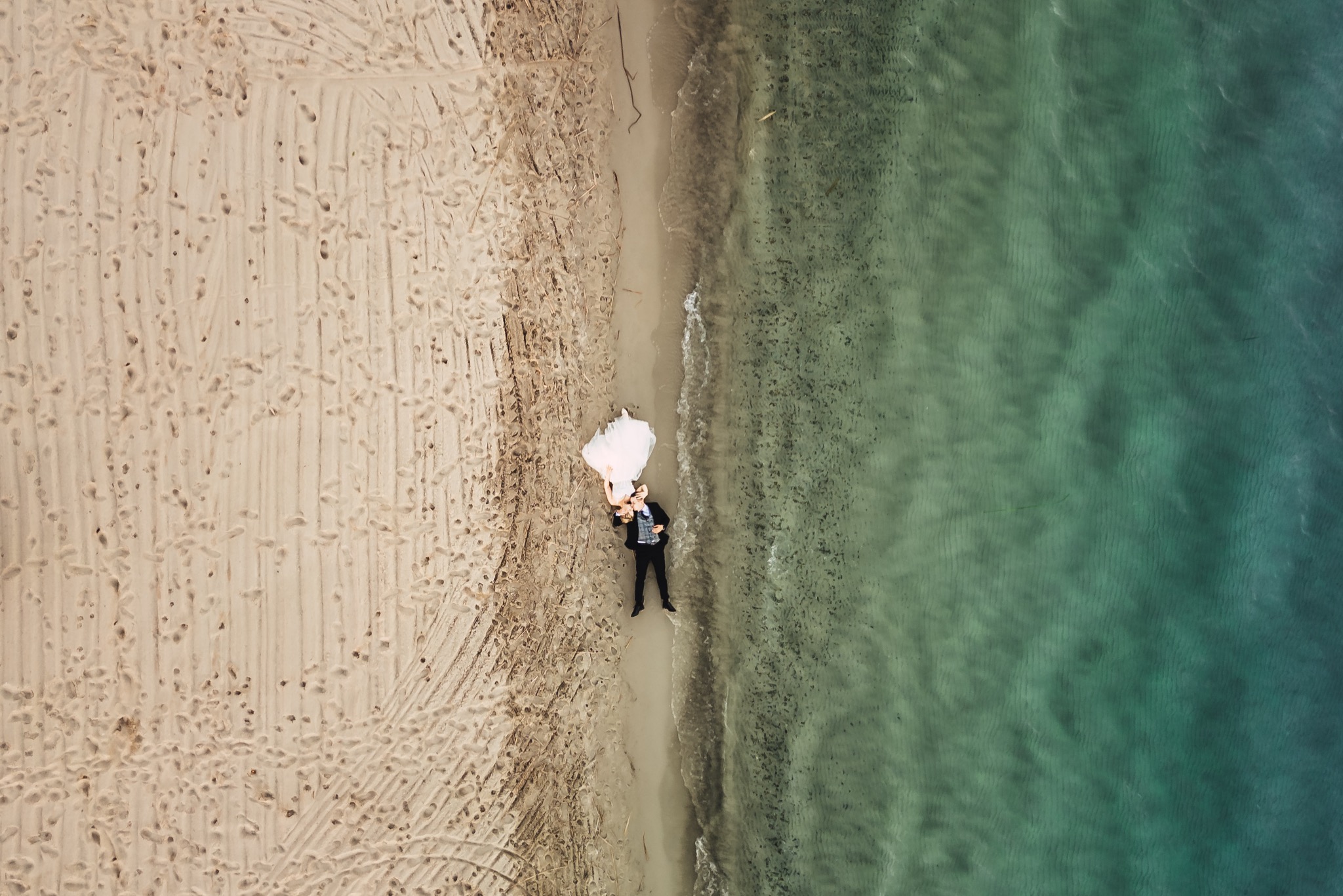 Para młoda leżąca na piaszczystej plaży, widok z lotu ptaka, granica piasku i turkusowej wody z widocznymi falami, ślady stóp na piasku.