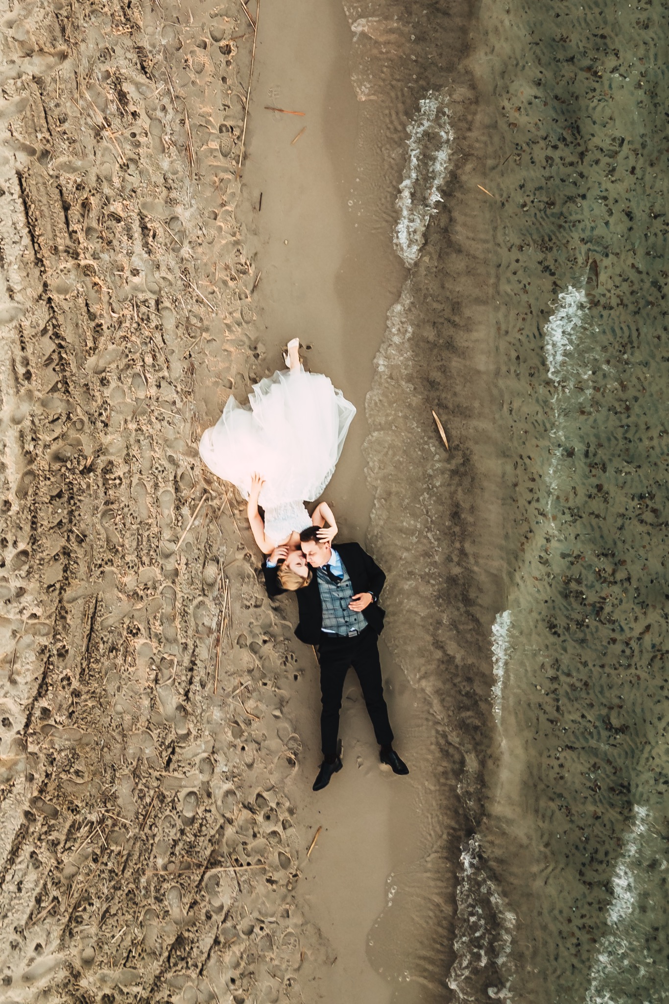 Nowożeńcy leżący na piasku plaży, widok z góry, panna młoda w białej sukni, pan młody w ciemnym garniturze, ślady stóp na piasku.