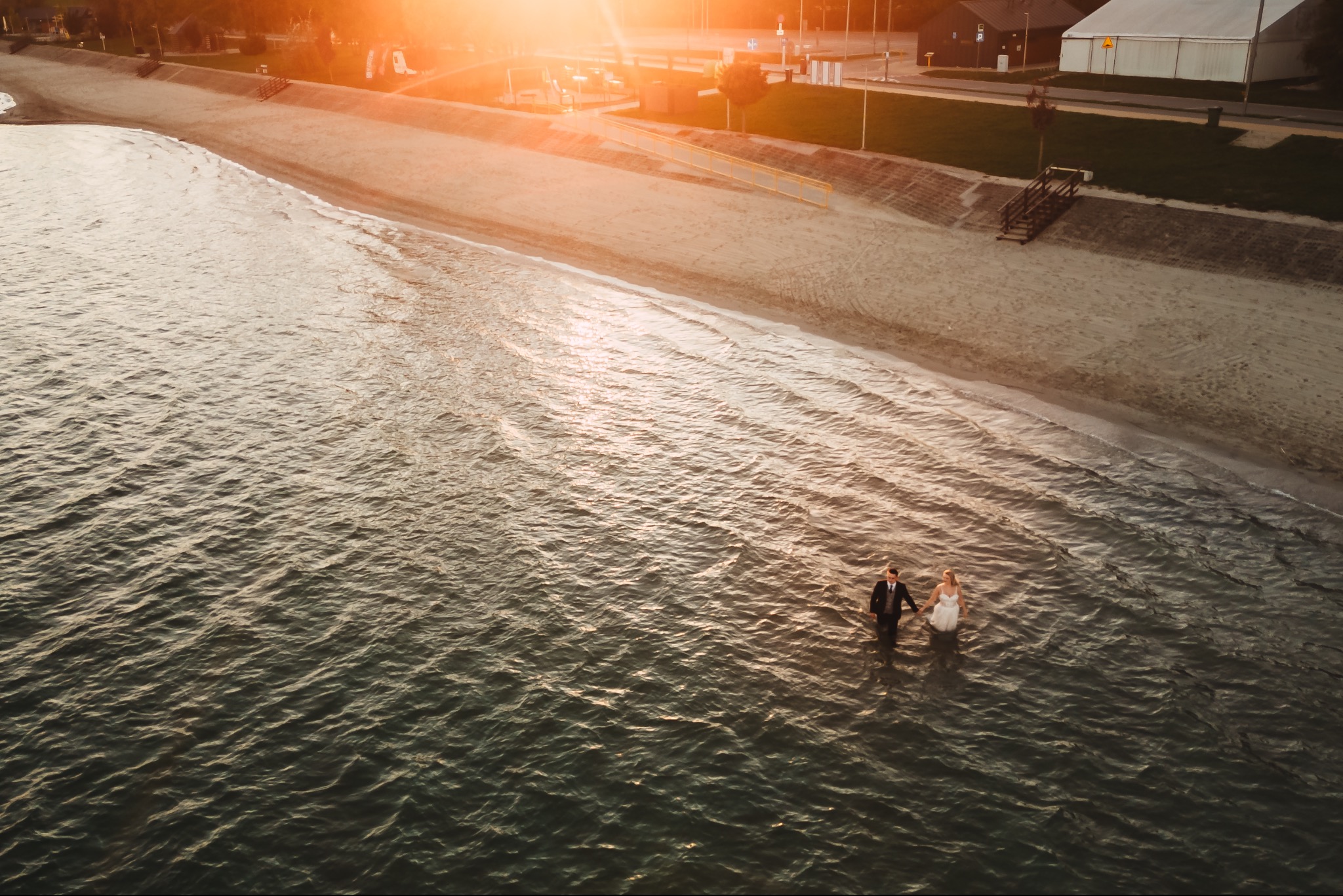 Para młoda trzymająca się za ręce, brodząca w płytkiej wodzie jeziora o zachodzie słońca, widok z lotu ptaka na brzeg i pobliską zabudowę.