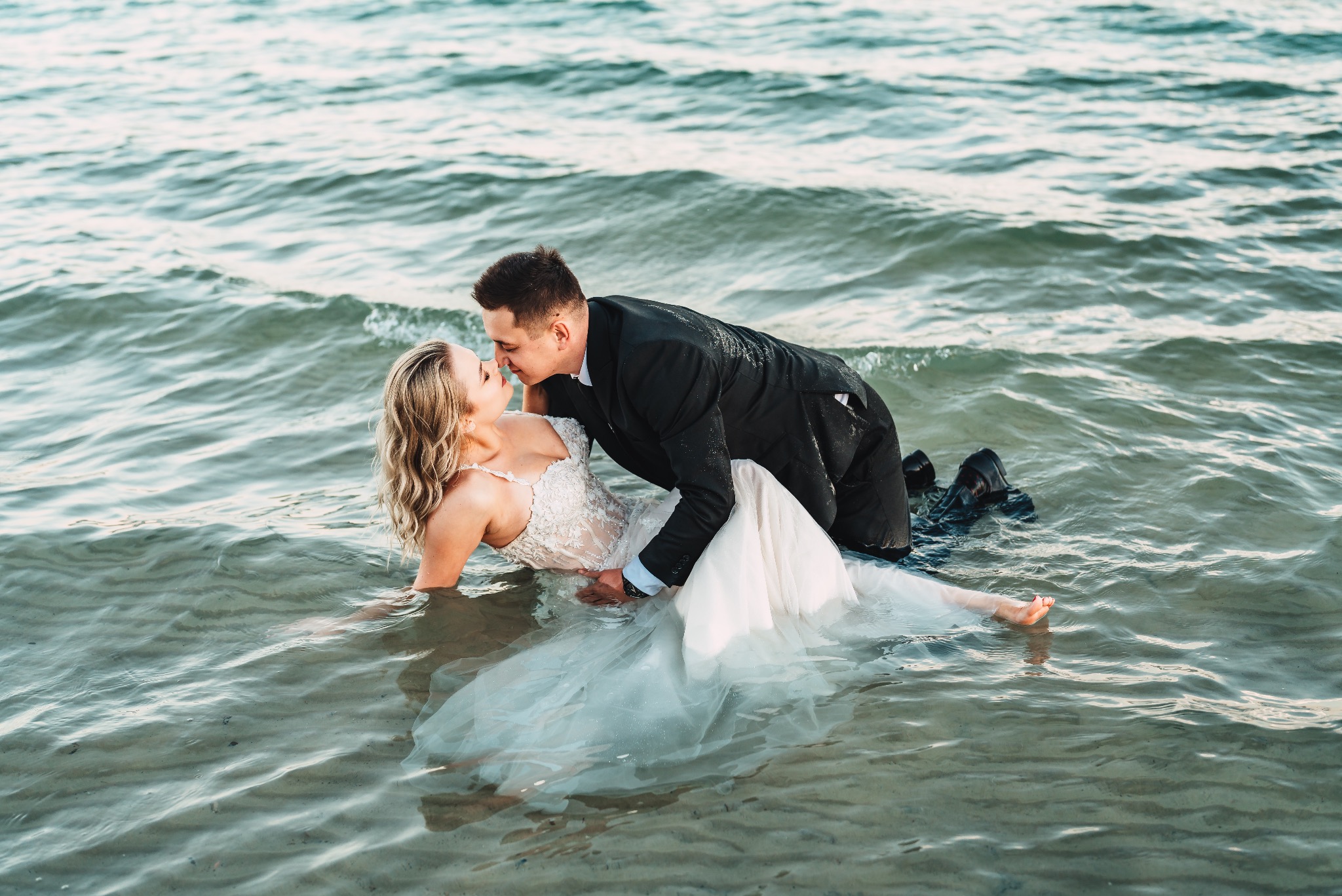 Para młoda w wodzie podczas pocałunku, panna młoda w koronkowej sukni, pan młody w czarnym garniturze, mokre ubrania, delikatne fale.