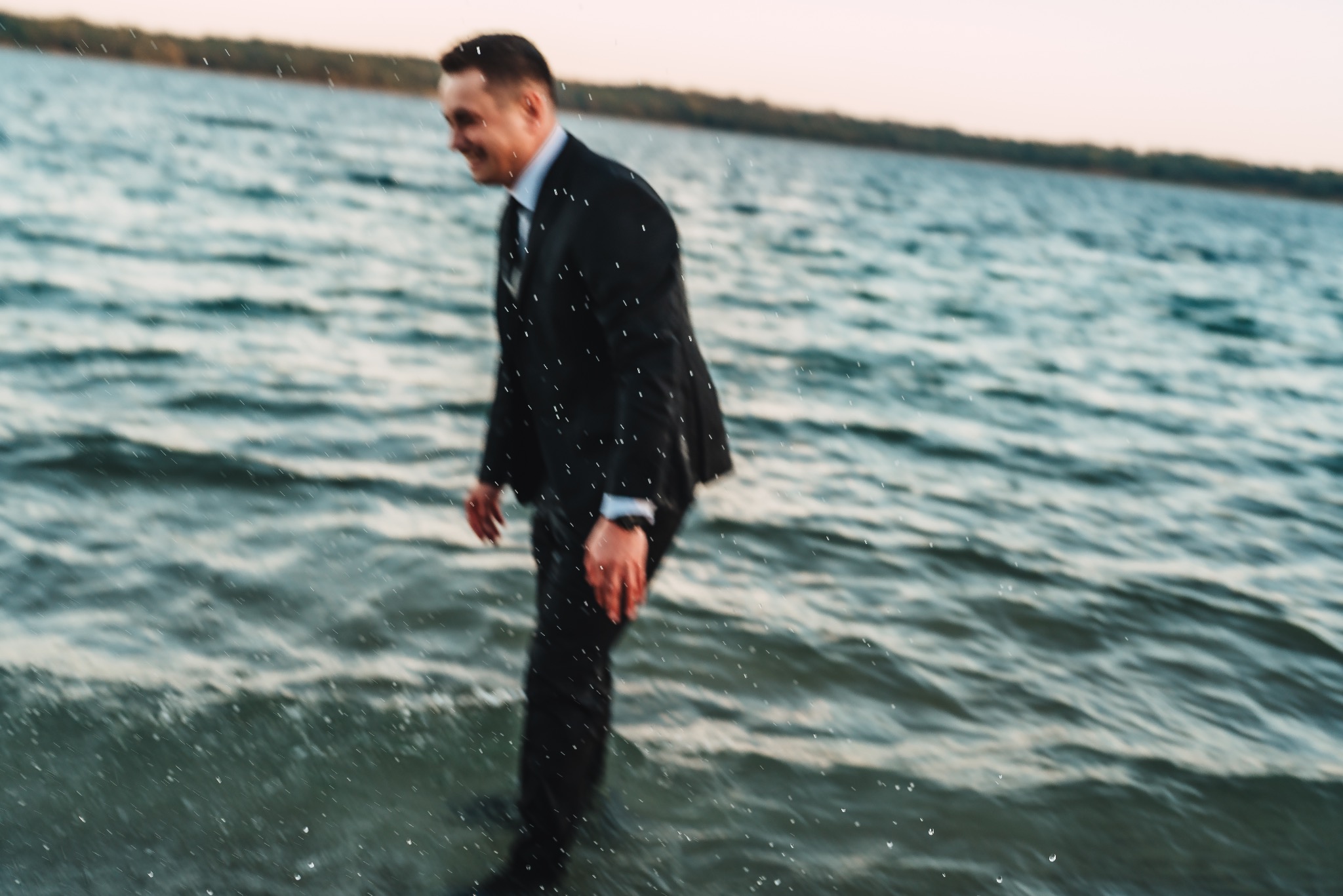 Mężczyzna w garniturze spaceruje po płytkiej wodzie jeziora, uśmiechając się, z zalesionym brzegiem w tle.