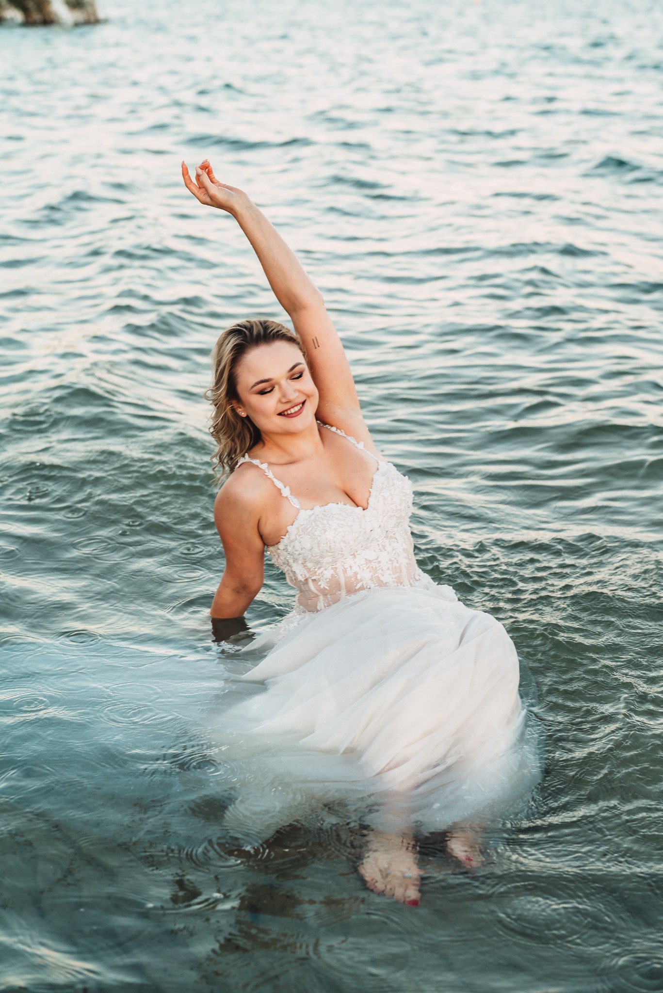 Uśmiechnięta panna młoda w koronkowej sukni, zanurzona w wodzie, z uniesioną ręką i widocznym tatuażem na ramieniu.