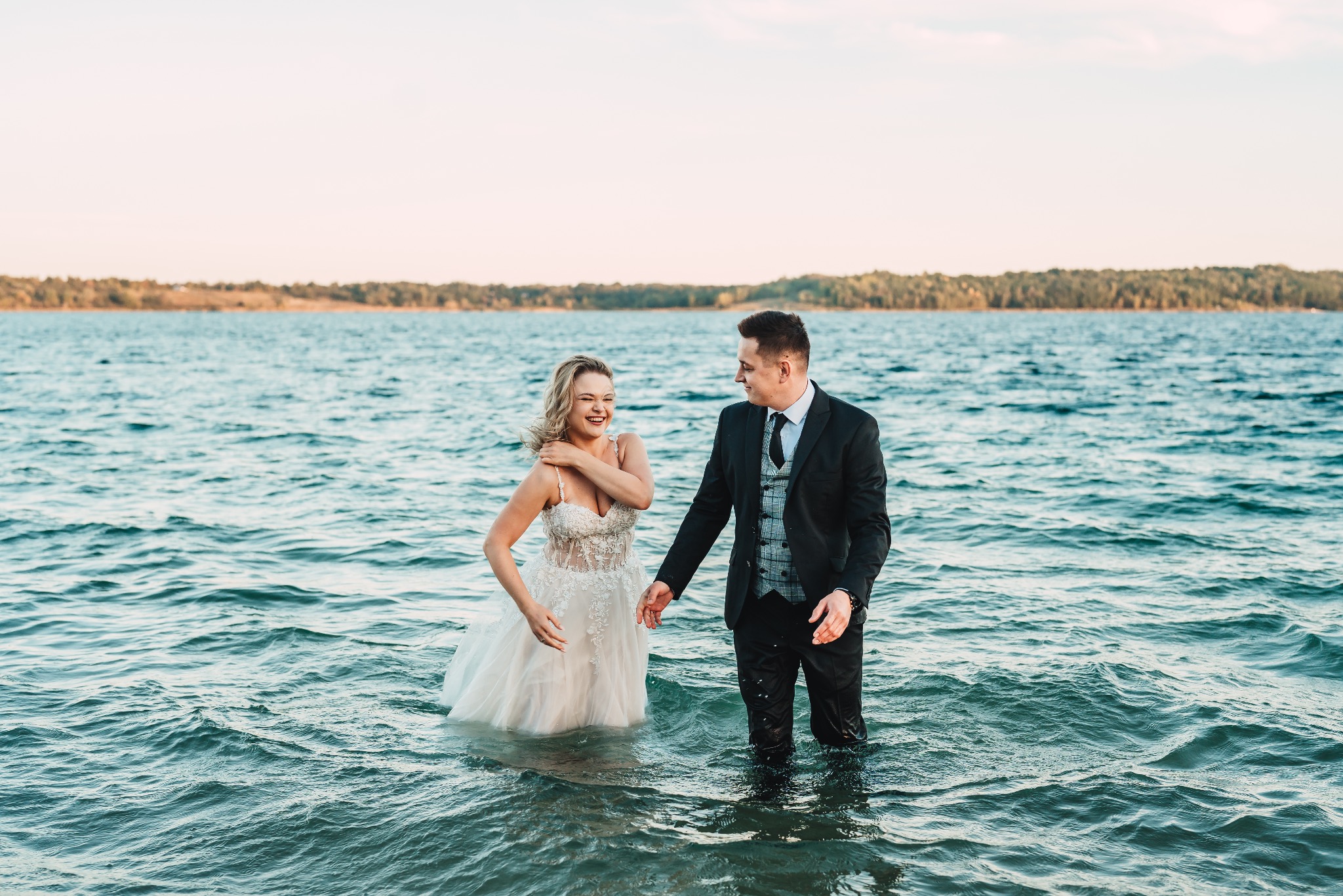 Uśmiechnięta panna młoda w koronkowej sukni i pan młody w garniturze stoją w wodzie jeziora podczas sesji ślubnej na tle zalesionego brzegu o zachodzie słońca.