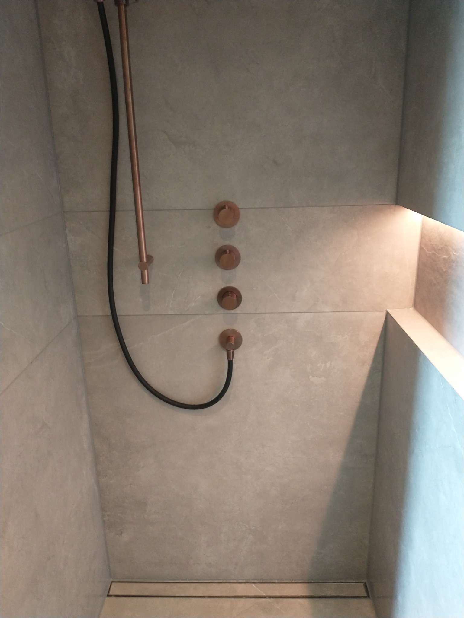 Nowoczesny prysznic z szarymi płytkami, miedzianą armaturą i odpływem liniowym w podłodze, oświetlenie LED w niszy.