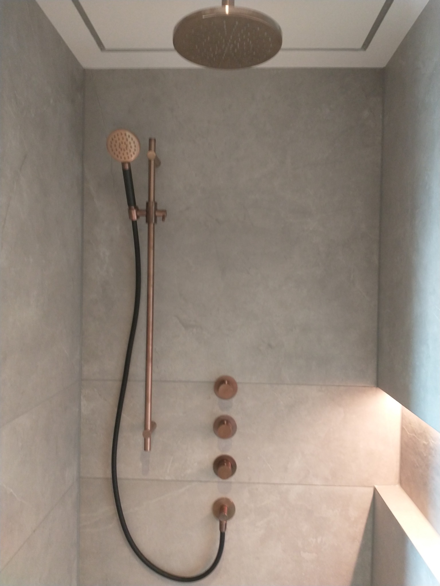 Nowoczesna kabina prysznicowa z deszczownicą i armaturą w kolorze miedzi, szare płytki imitujące kamień, minimalistyczne oświetlenie wnęki.