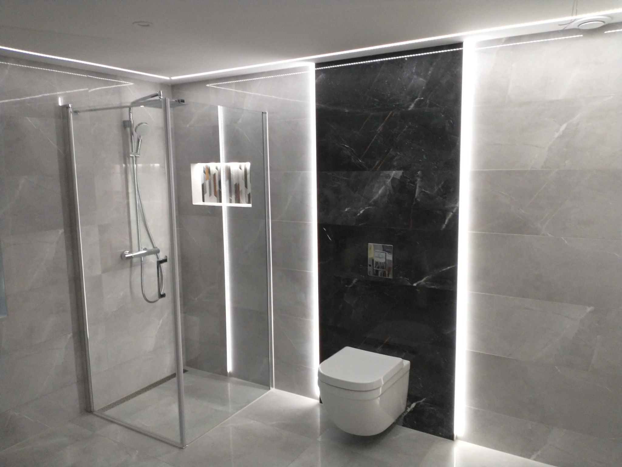 Nowoczesna łazienka z kabiną prysznicową typu walk-in, podwieszaną toaletą i oświetleniem LED wzdłuż ścian z szarych i czarnych marmurkowych płytek.