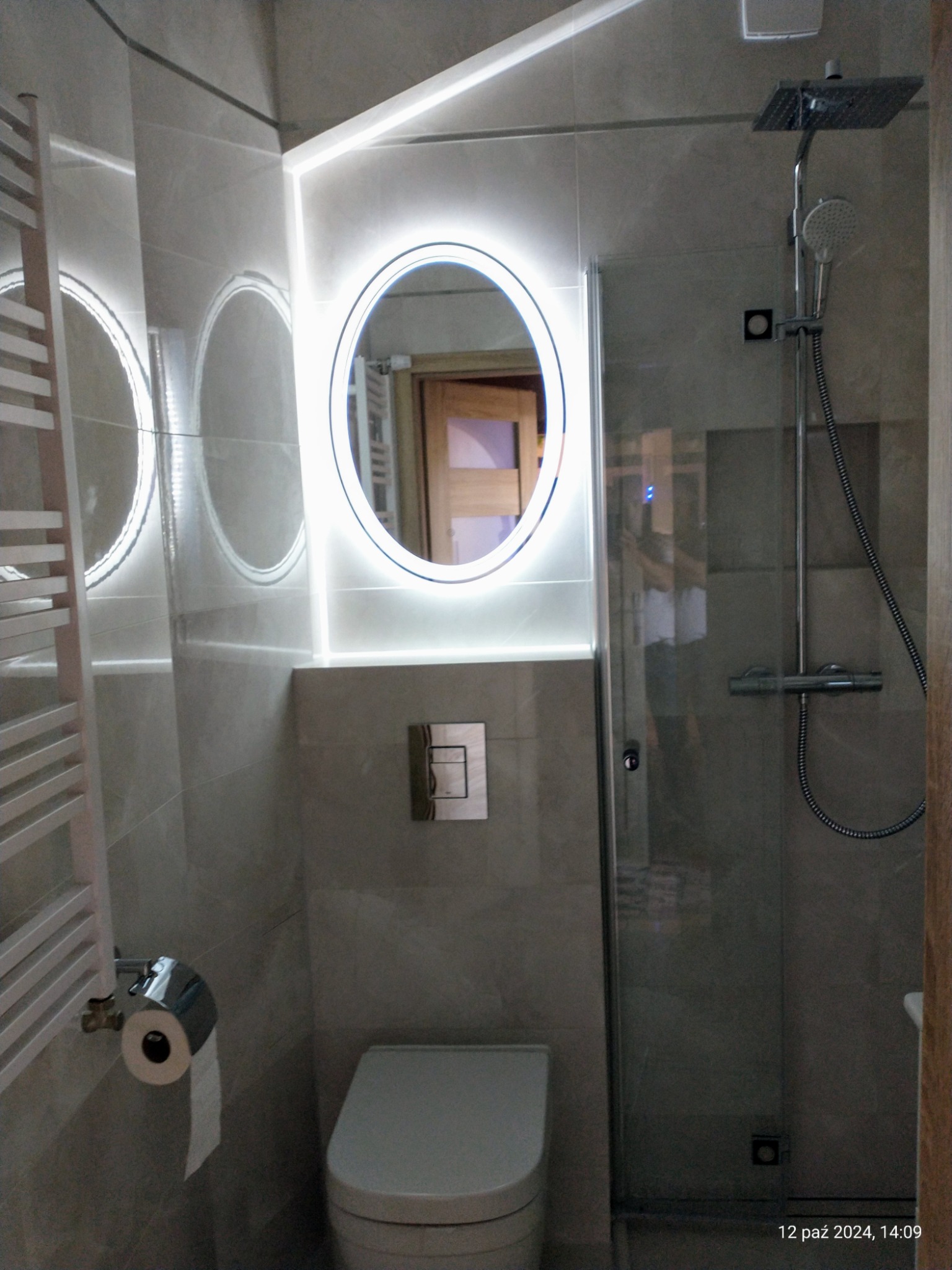 Nowoczesna łazienka z podświetlanym okrągłym lustrem, kabiną prysznicową typu walk-in i grzejnikiem drabinkowym w odcieniach szarości.