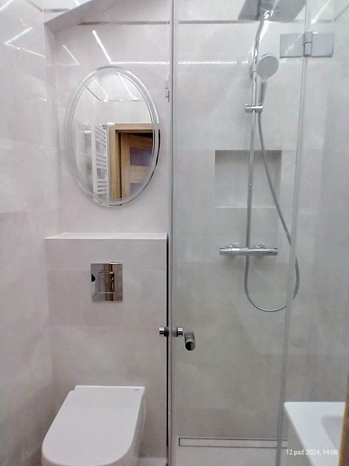 Nowoczesna łazienka z białymi płytkami, kabiną prysznicową ze szklanymi drzwiami, owalnym lustrem i podtynkową toaletą z chromowanym przyciskiem spłukiwania.