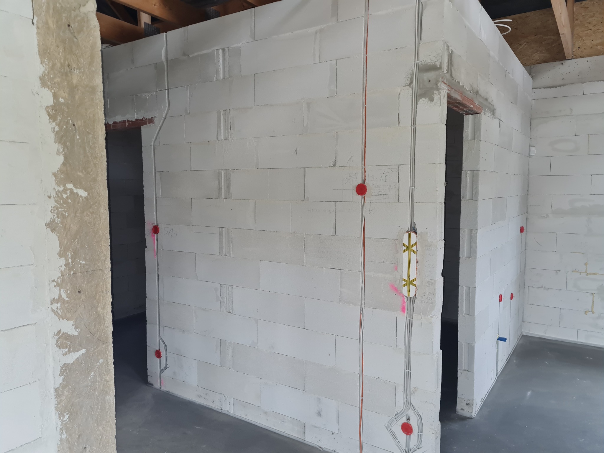 Wnętrze w trakcie budowy z widocznymi przewodami elektrycznymi w ścianach z betonu komórkowego, czerwone puszki instalacyjne i przewody poprowadzone w bruzdach.
