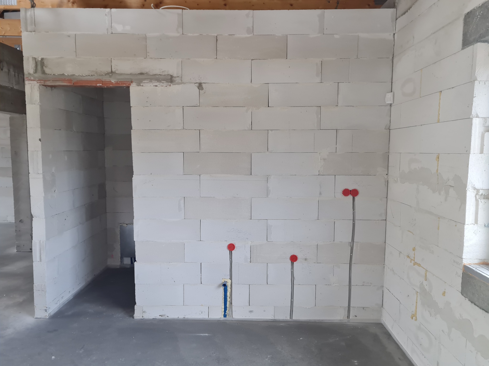 Wewnętrzna ściana z betonu komórkowego w trakcie budowy, widoczne wystające rury instalacji elektrycznej zabezpieczone czerwonymi zaślepkami oraz niebieska rura instalacji wodnej, surowy stan...