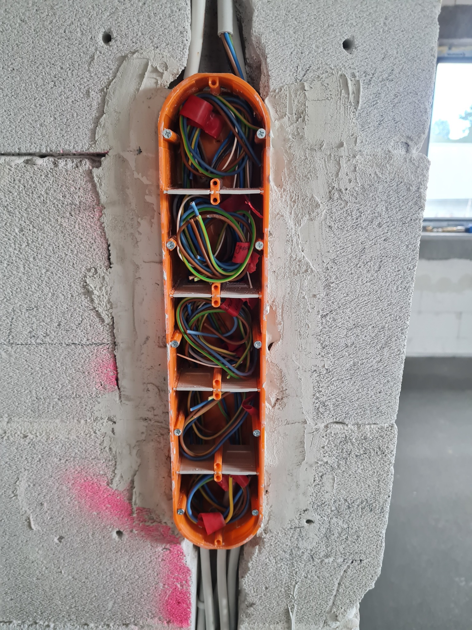 Pomarańczowa puszka elektryczna podtynkowa, zamontowana w surowej ścianie z betonu komórkowego, z widocznymi przewodami w różnych kolorach, zabezpieczonymi czerwonymi złączkami. Widoczne ślady...