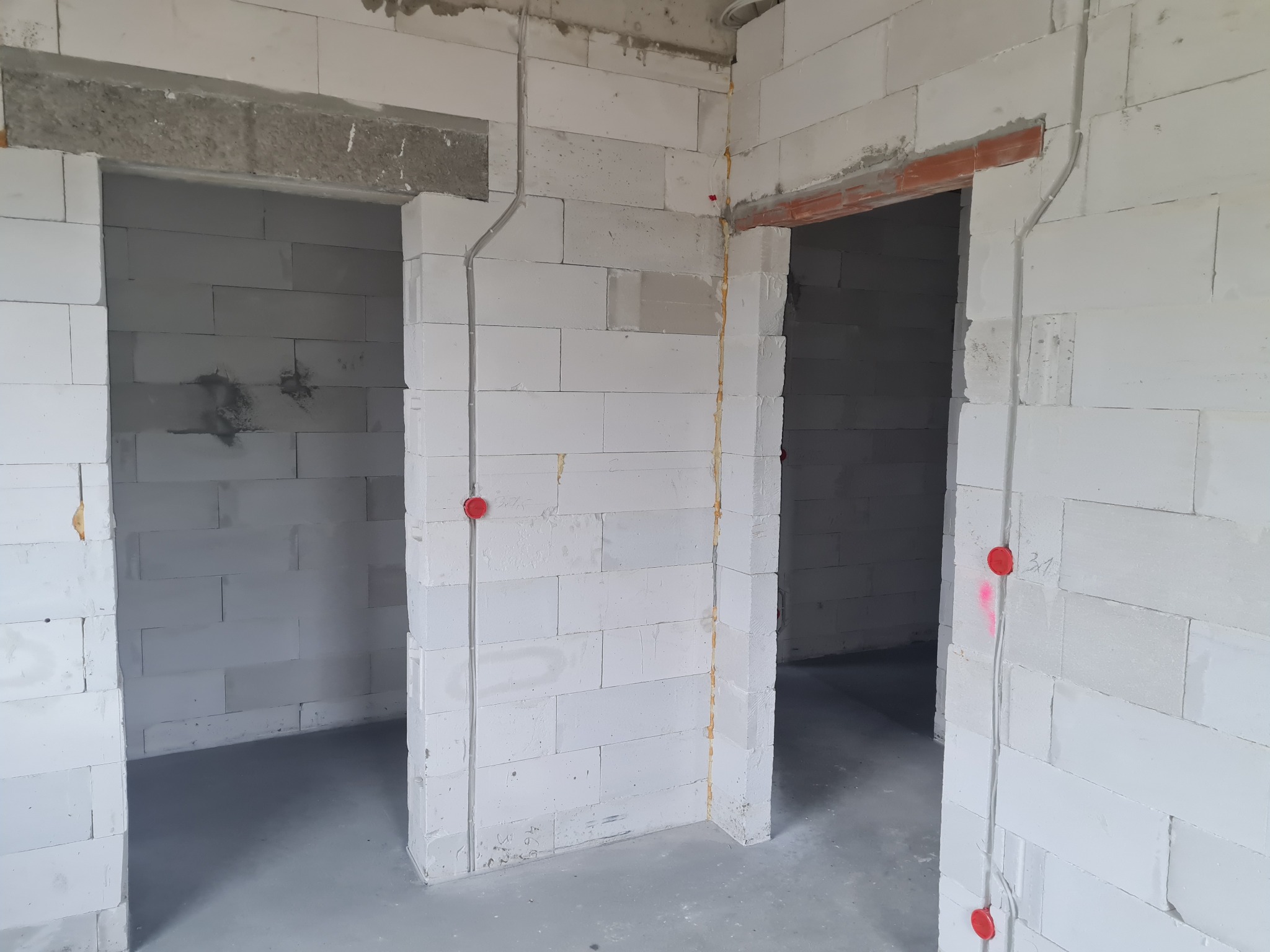 Wewnętrzne ściany z betonu komórkowego w trakcie budowy, widoczne peszle z przewodami elektrycznymi zabezpieczone czerwonymi zaślepkami, surowy stan wykończenia.