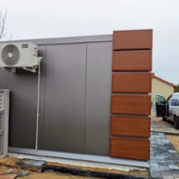 Zewnętrzna jednostka klimatyzatora AUX zamontowana na szarej ścianie budynku z brązowymi panelami dekoracyjnymi, obok zaparkowany biały samoch&oacute;d dostawczy z reklamą firmy klimatyzacyjnej.