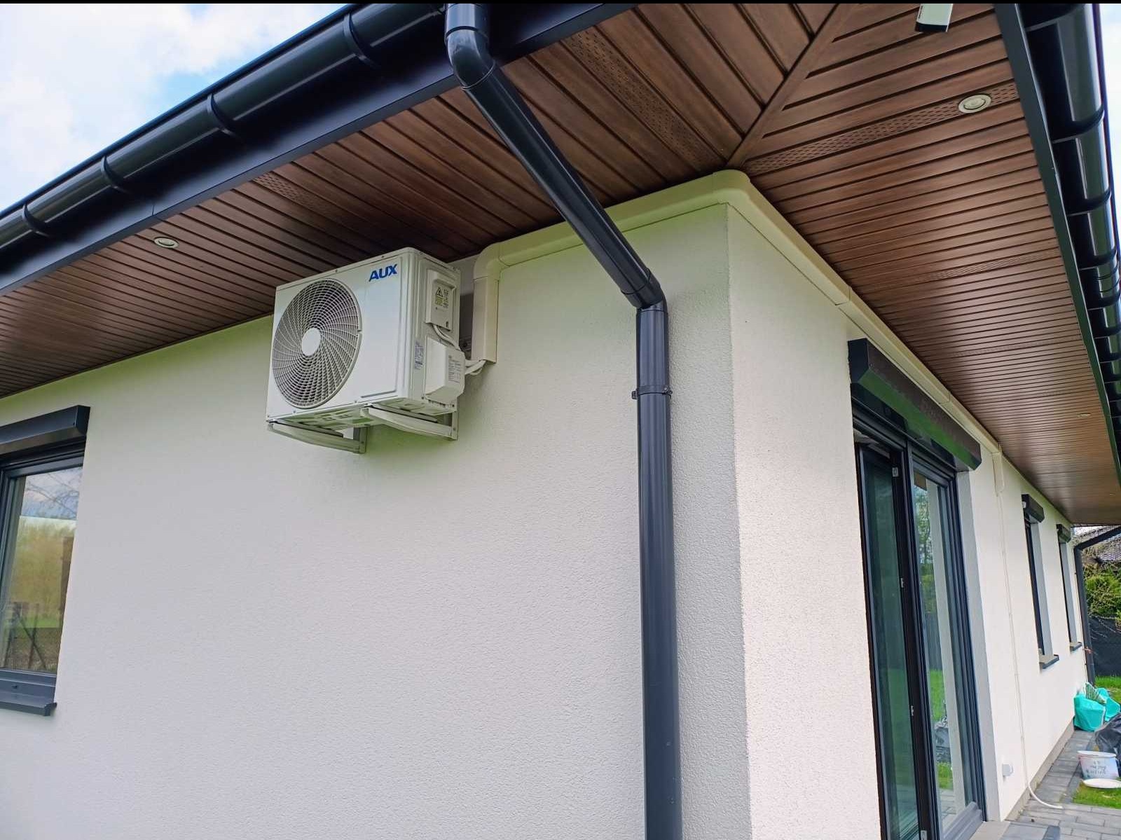 Zewnętrzna jednostka klimatyzacji AUX zamontowana na elewacji domu z szarymi rynnami i drewnianą podbitką dachową.