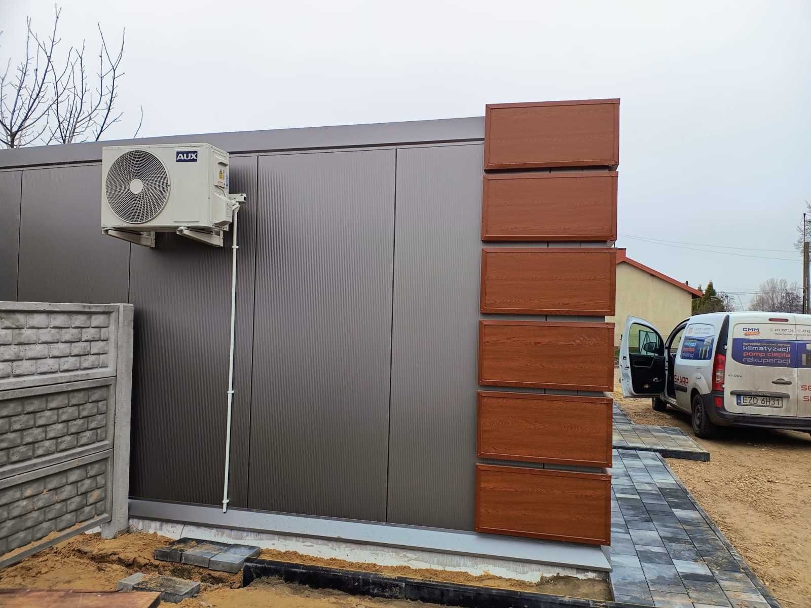 Zewnętrzna jednostka klimatyzatora AUX zamontowana na szarej ścianie budynku z brązowymi panelami dekoracyjnymi, obok zaparkowany biały samochód dostawczy z reklamą firmy klimatyzacyjnej.
