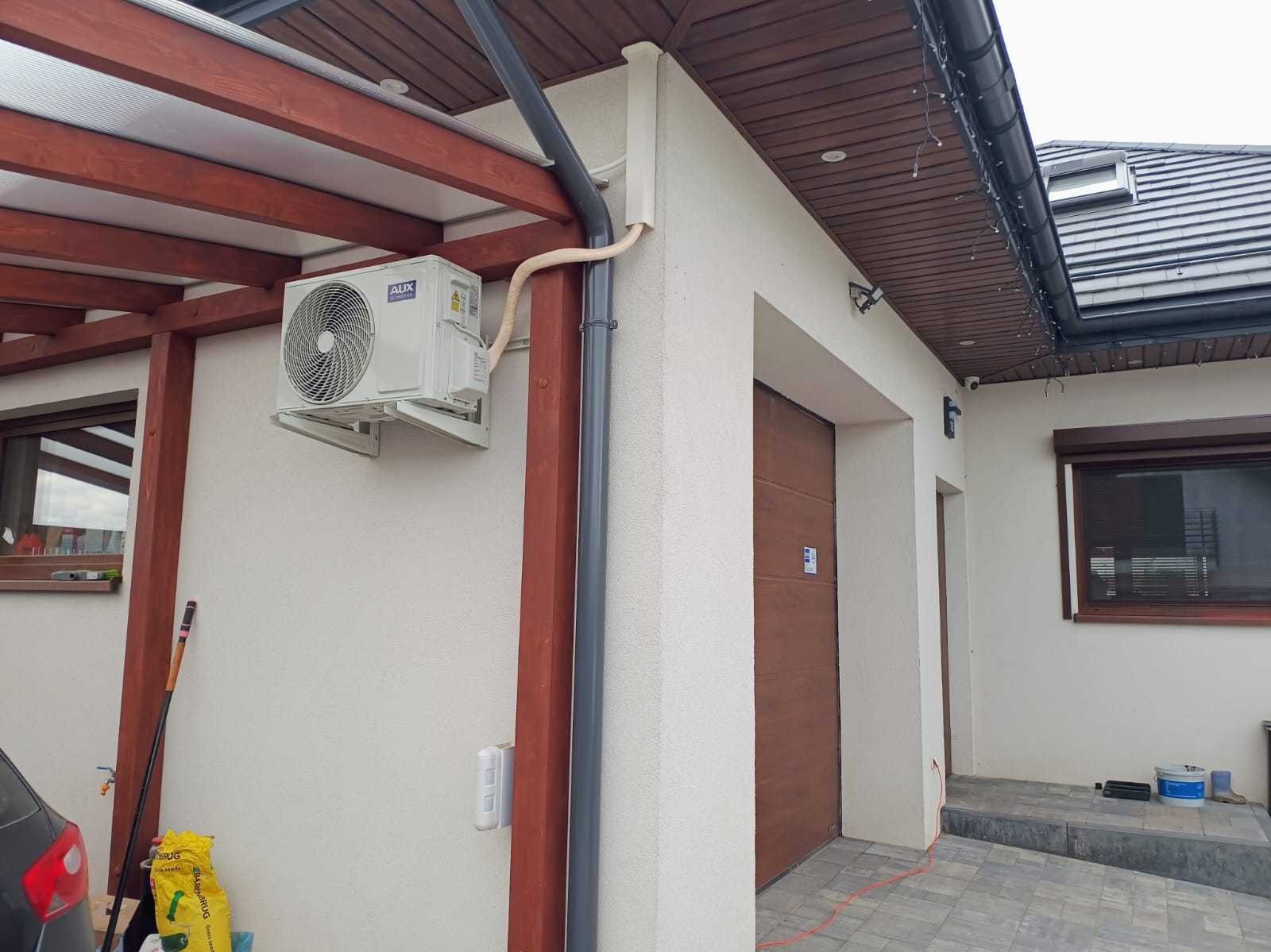 Zewnętrzna jednostka klimatyzatora marki AUX zamontowana na ścianie budynku z drewnianą konstrukcją zadaszenia w tle.