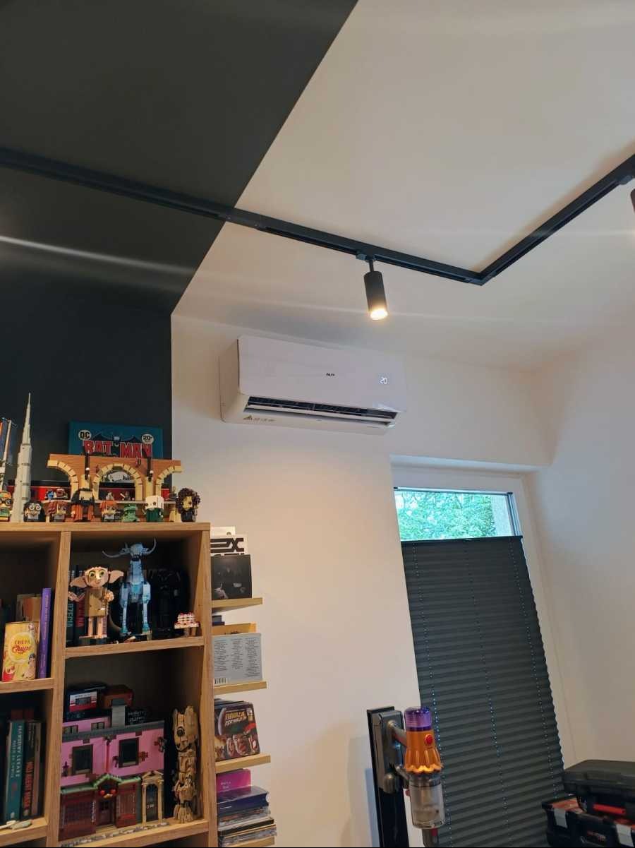 Zamontowana biała klimatyzacja AUX w pokoju z czarno-białym sufitem, oświetleniem szynowym i regałem z zabawkami.