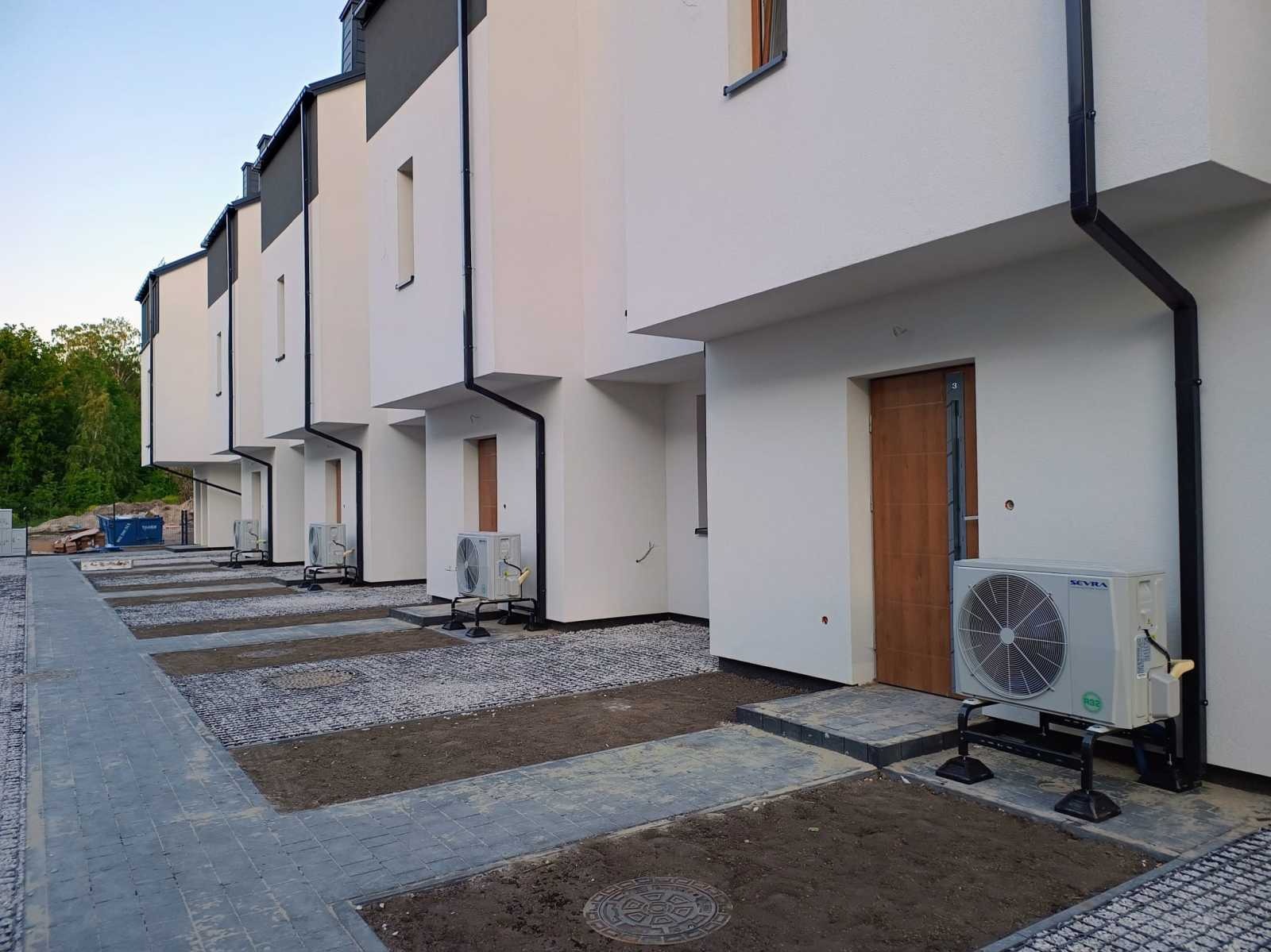 Zewnętrzne jednostki klimatyzacji zamontowane na wspornikach przy szeregowych domach jednorodzinnych z białymi elewacjami i brązowymi drzwiami wejściowymi, widok z poziomu gruntu na nowo...