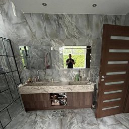 Łukasz - Wykończona łazienka z marmurowymi płytkami na ścianach i podłodze, drewnianą szafką pod umywalką z marmurowym blatem i dużym lustrem, w którym odbija się osoba robiąca zdjęcie.